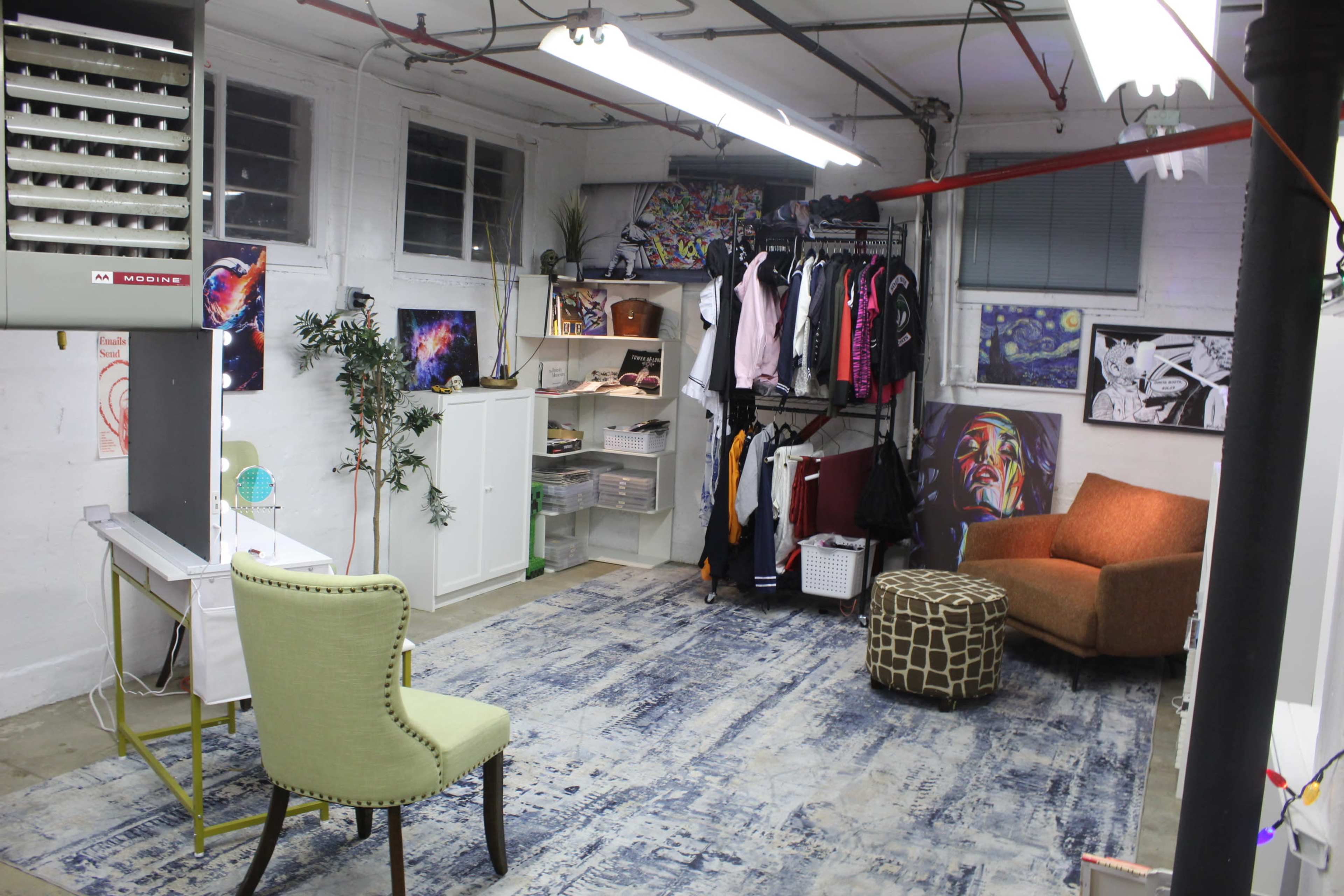 The image depicts a spacious basement studio with a clothing rack, storage shelves, an armchair, and various artworks displayed on the walls.