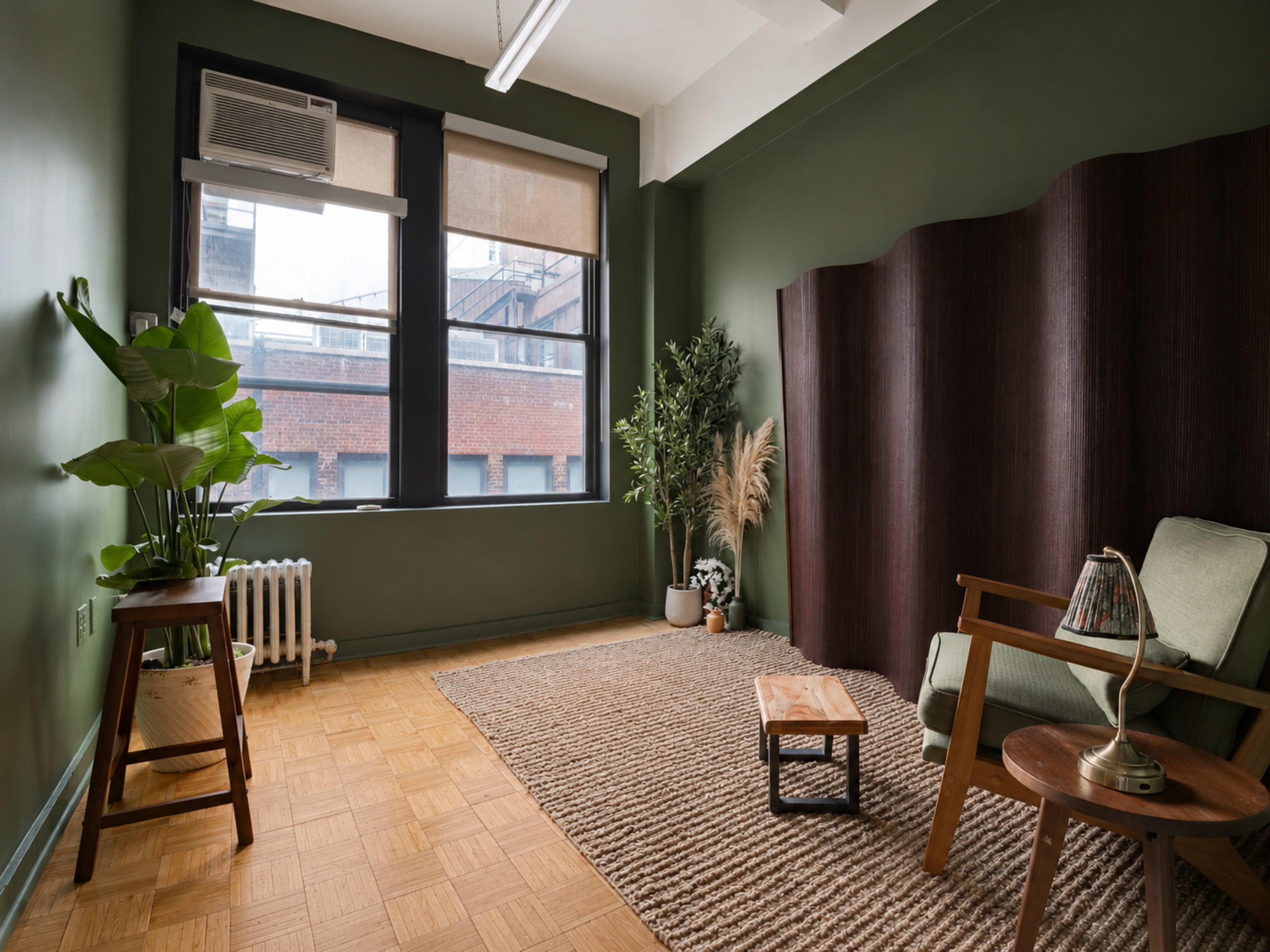 The image shows a cozy interior room with olive green walls, a wooden floor, a large plant, a small wooden table, and a comfortable chair against a partition.