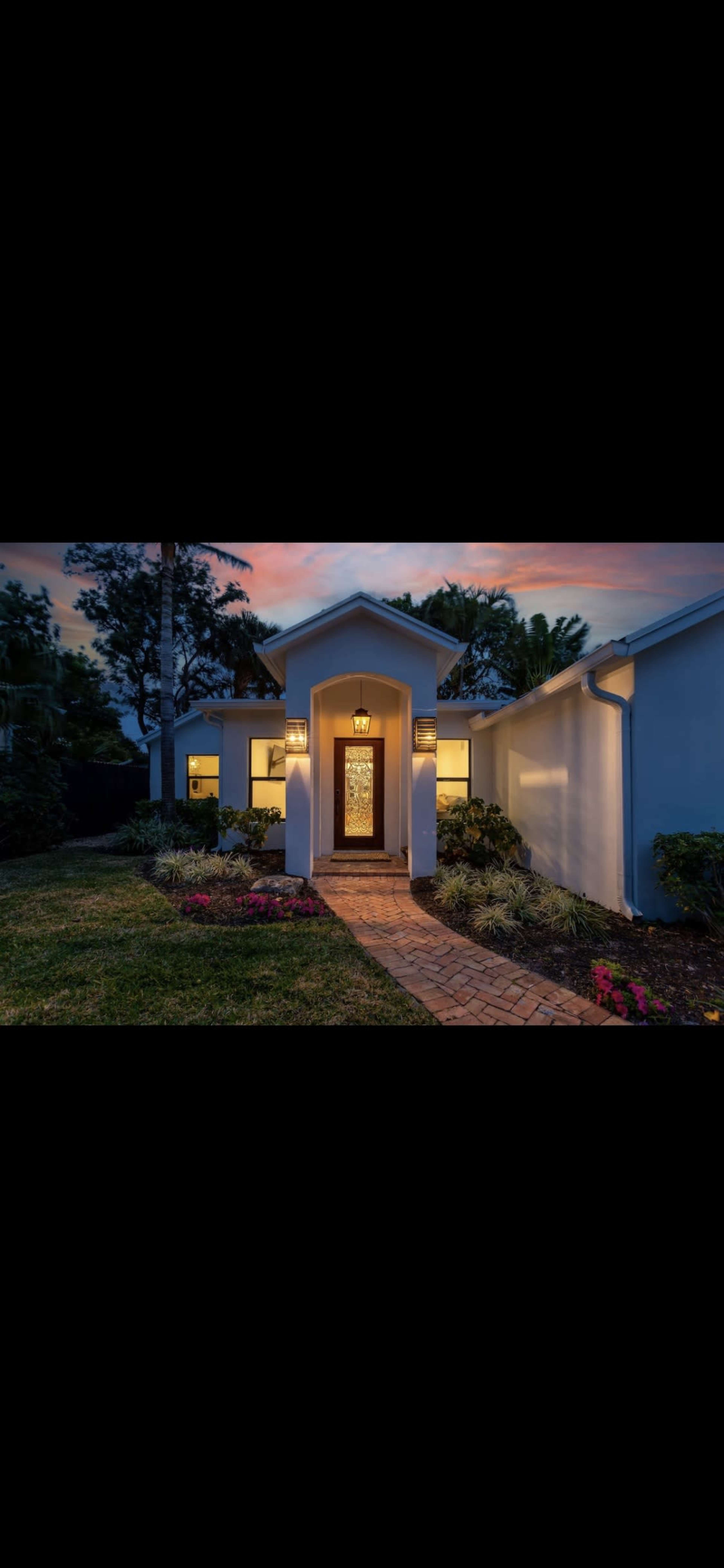 The image shows a modern home with a lighted entryway surrounded by landscaped gardens and a brick pathway, under a vibrant sky at dusk.