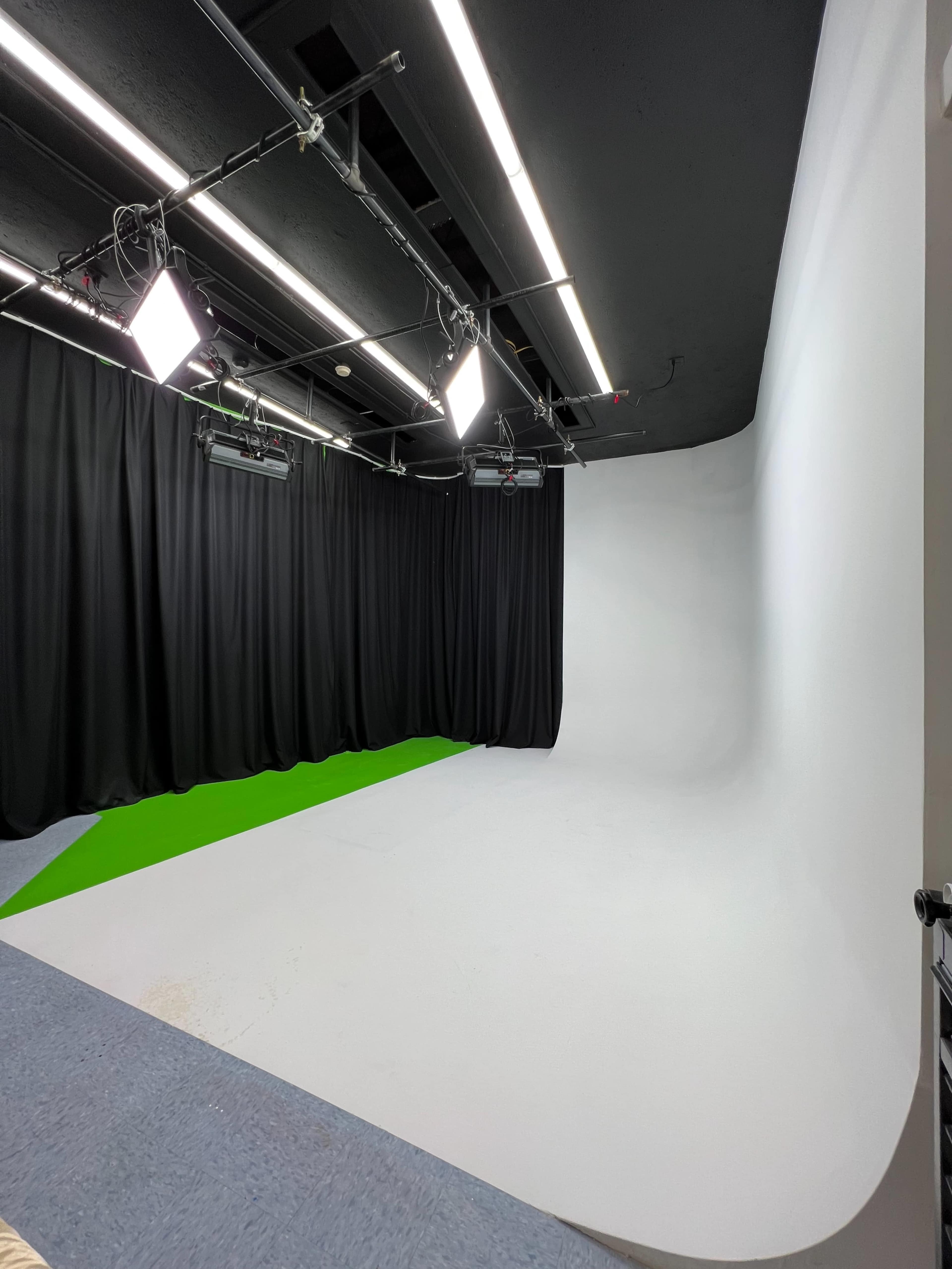 The image shows a studio space with a white seamless backdrop, green floor section, and overhead lights, surrounded by black curtains.