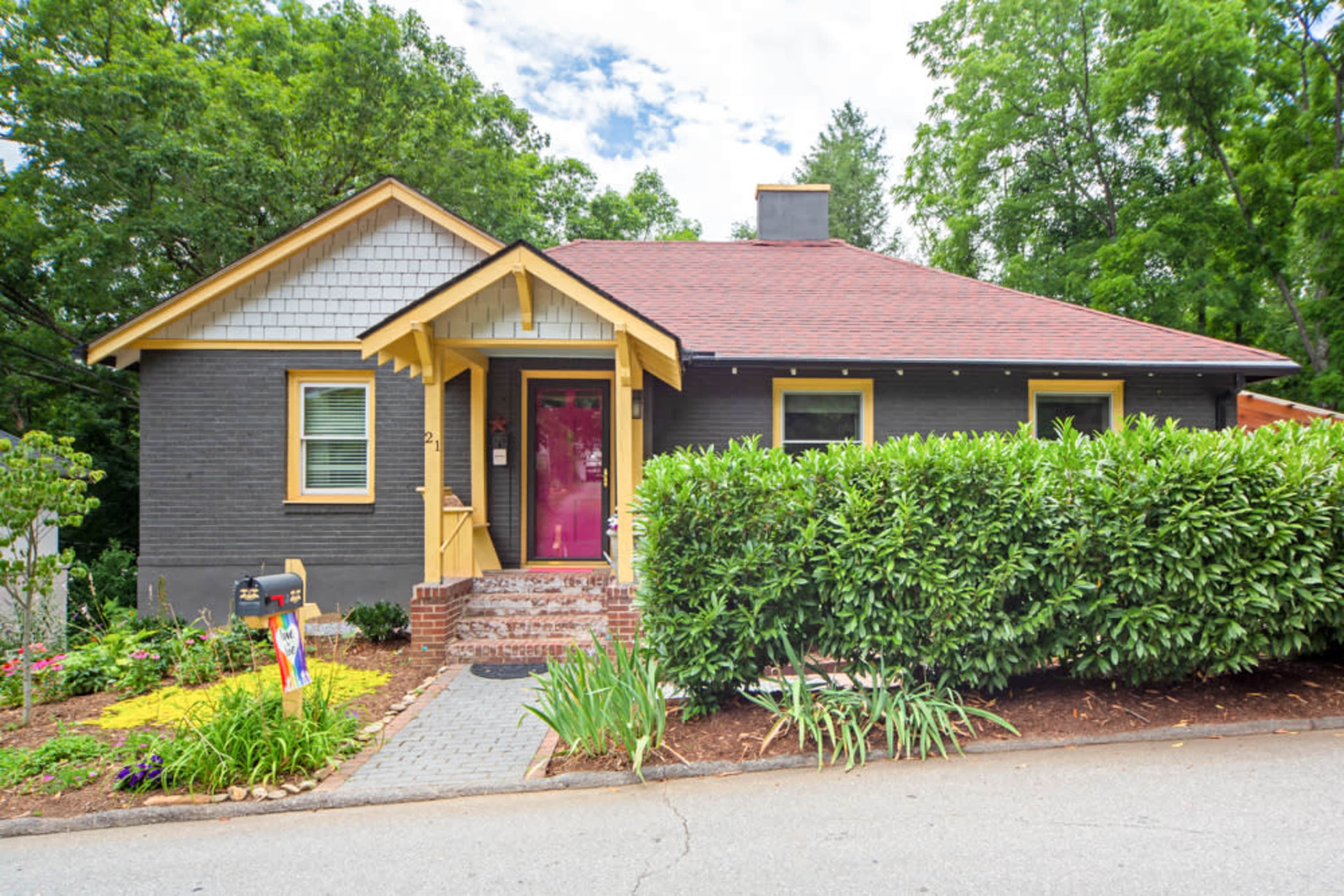 A one-story gray house with a red roof and a bright pink door is surrounded by green shrubs and a landscaped front yard.