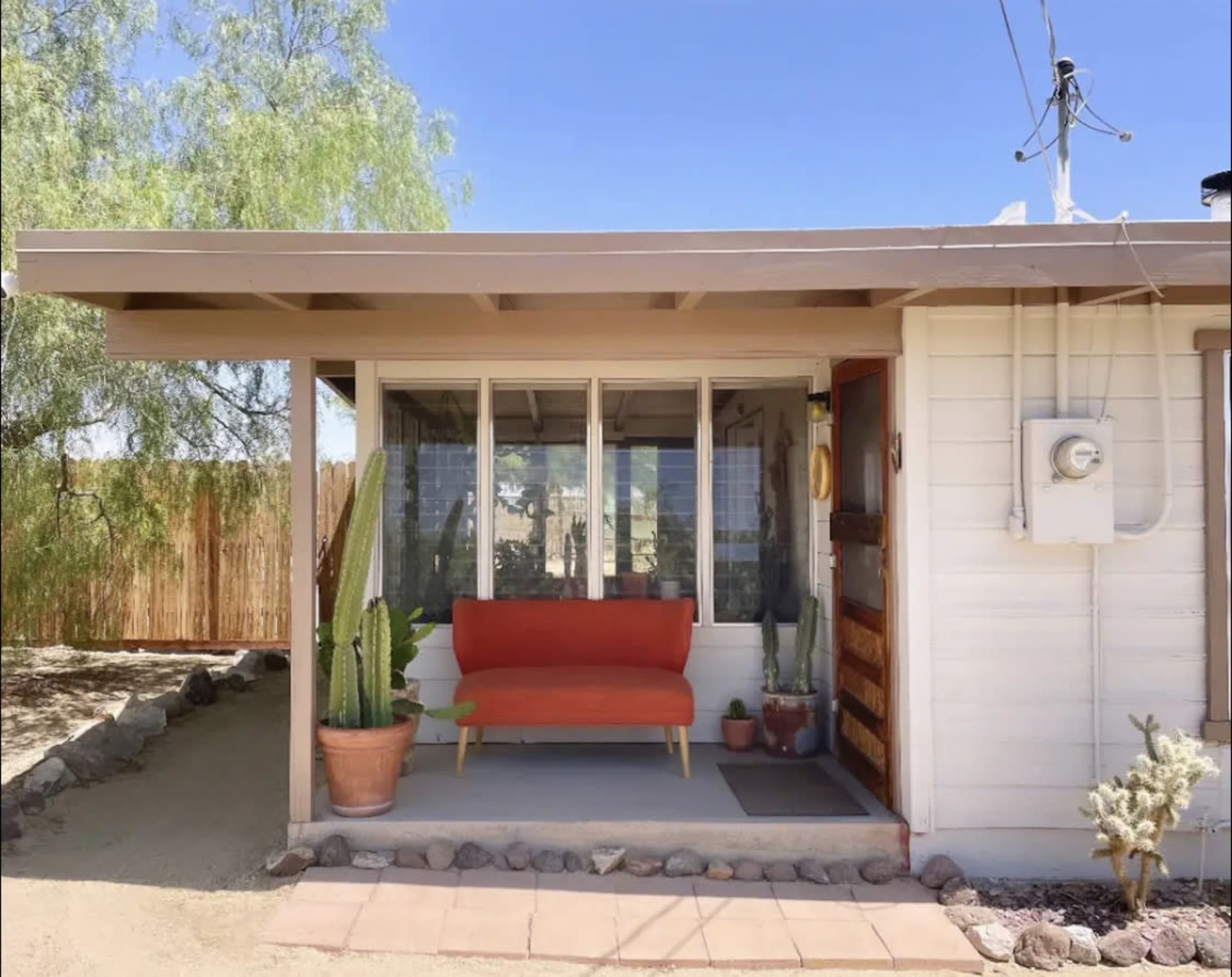 A bright red sofa sits on a porch in front of a small house surrounded by desert landscaping and tall cacti.