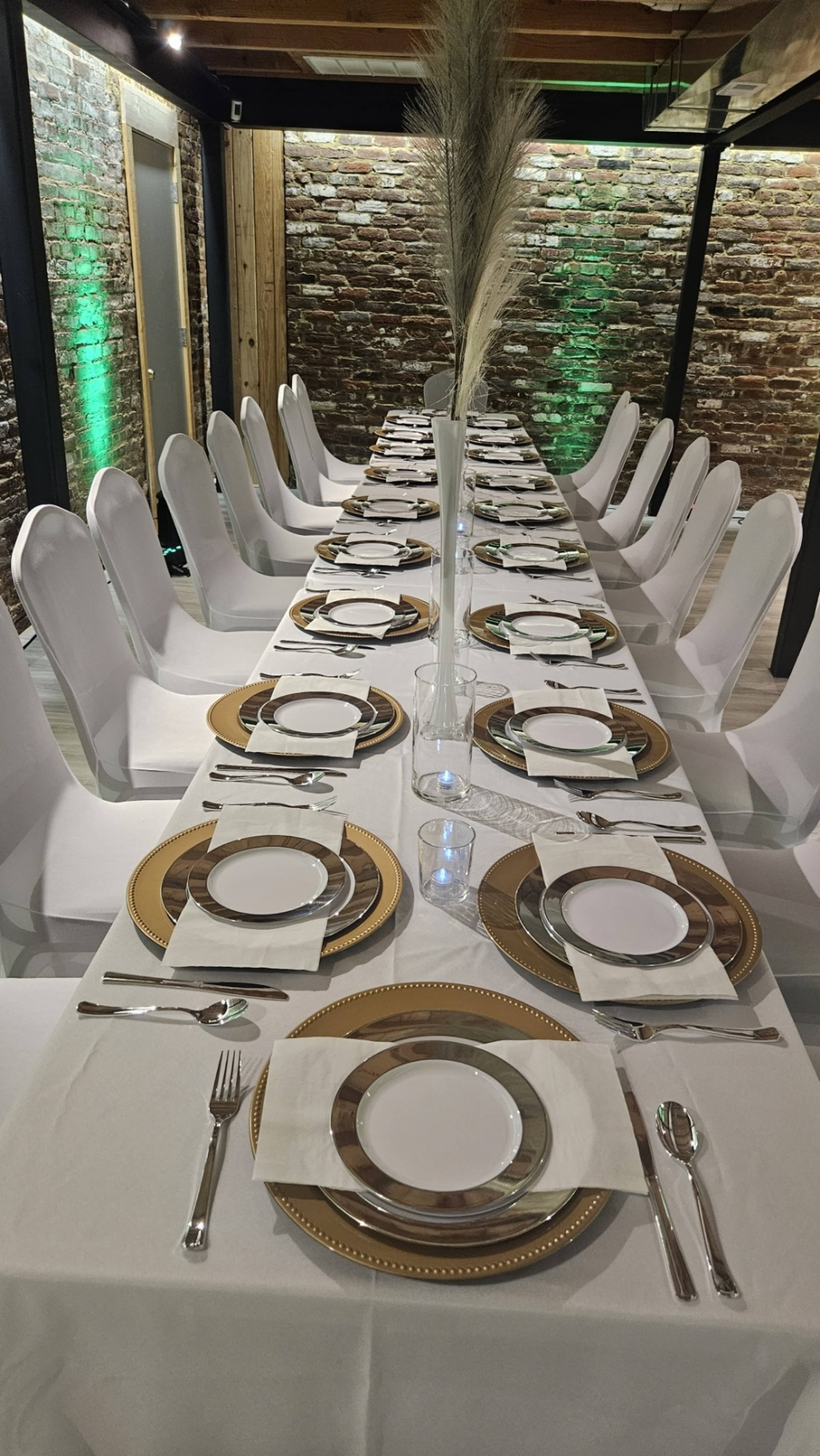 A long dining table is elegantly set with white and gold tableware in a well-lit indoor space featuring exposed brick walls.