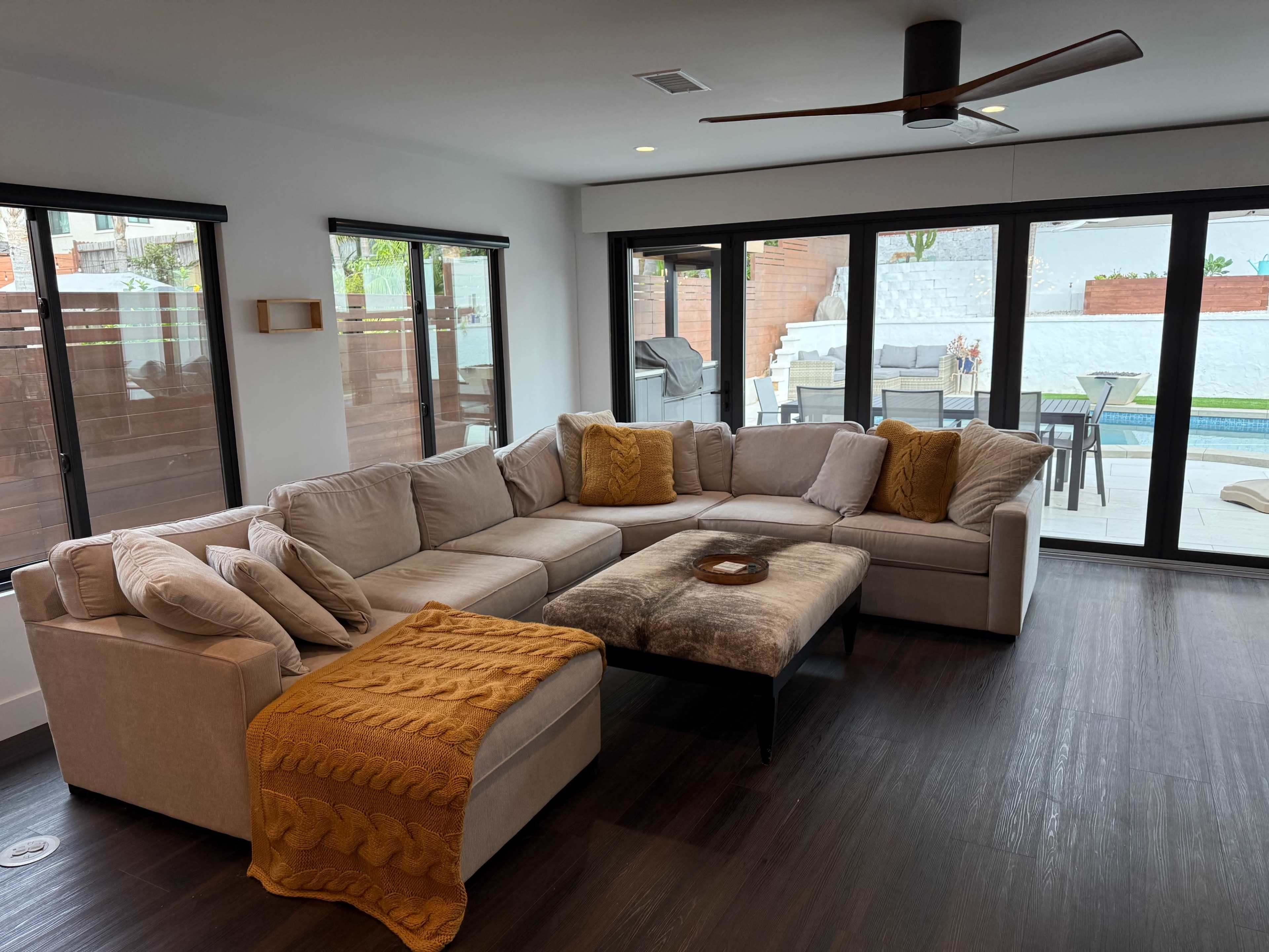 The image shows a modern living room with a large sectional sofa, a coffee table, and large windows overlooking an outdoor patio with a pool.