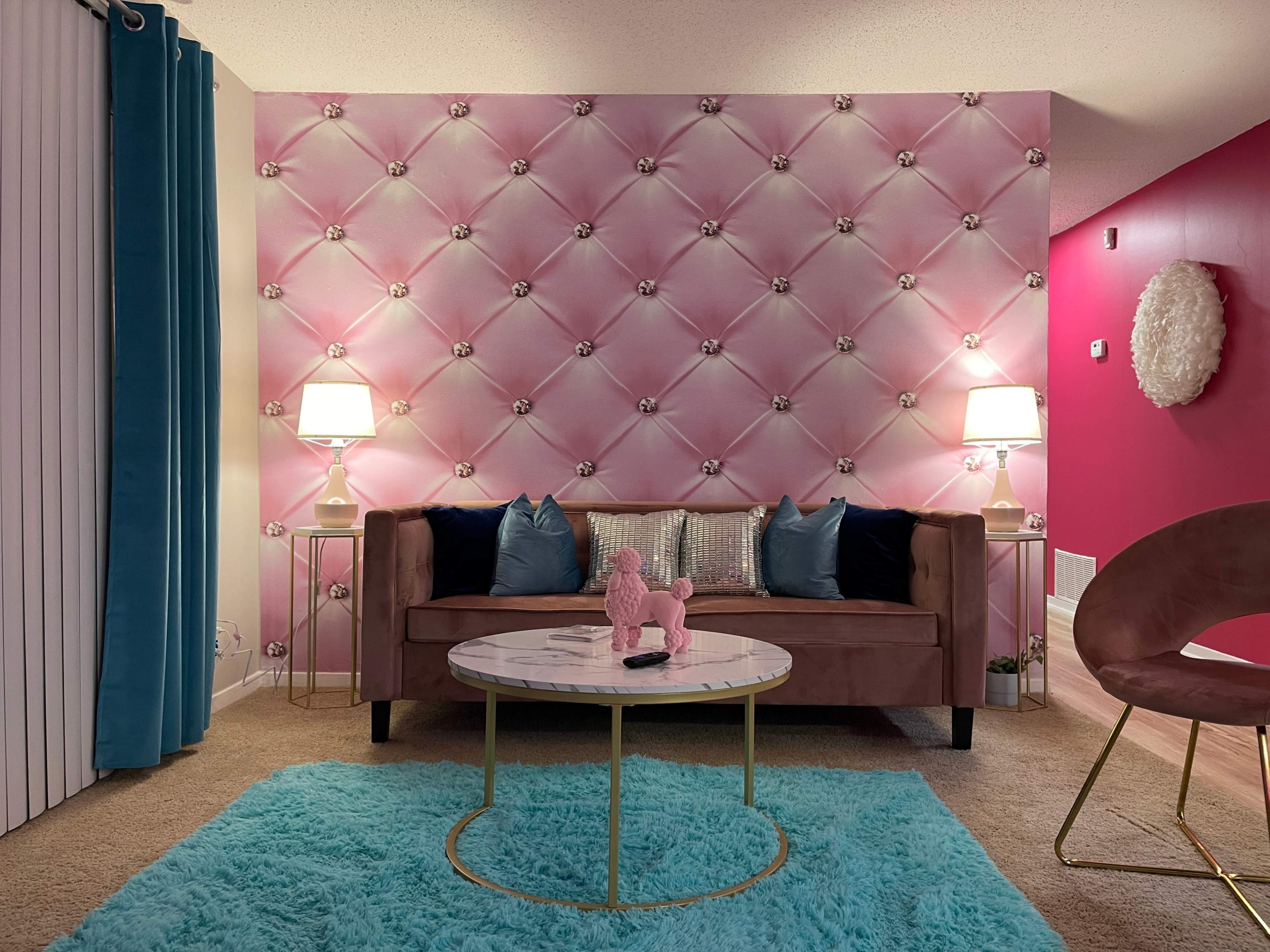 A living room features a pink tufted wall, a brown sofa with decorative cushions, a round coffee table, and a turquoise rug.