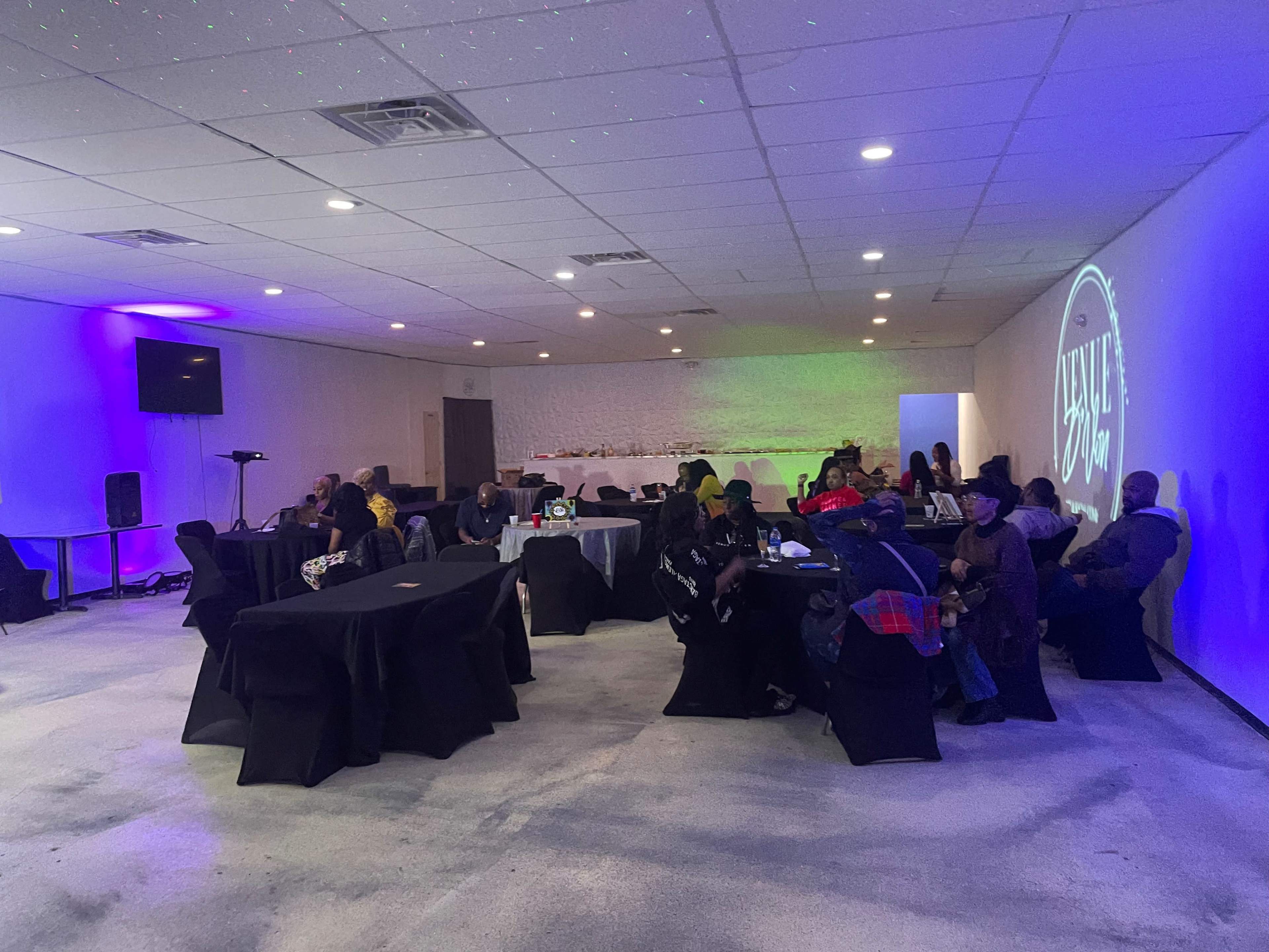 A group of people sits at tables in a dimly lit room with purple lighting and a projected logo on the wall.