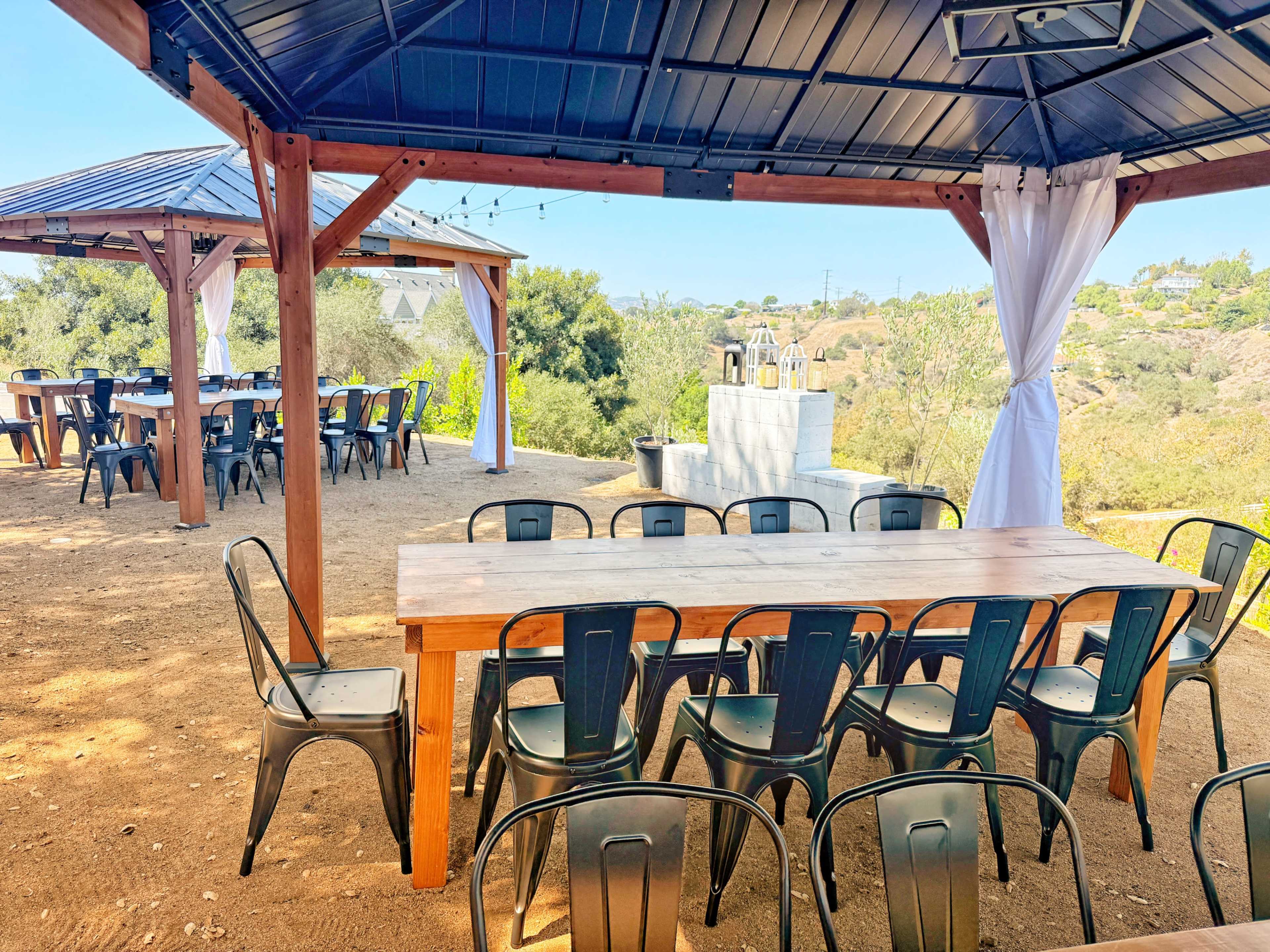 The image shows two wooden tables with metal chairs arranged under a gazebo in an outdoor setting surrounded by greenery.