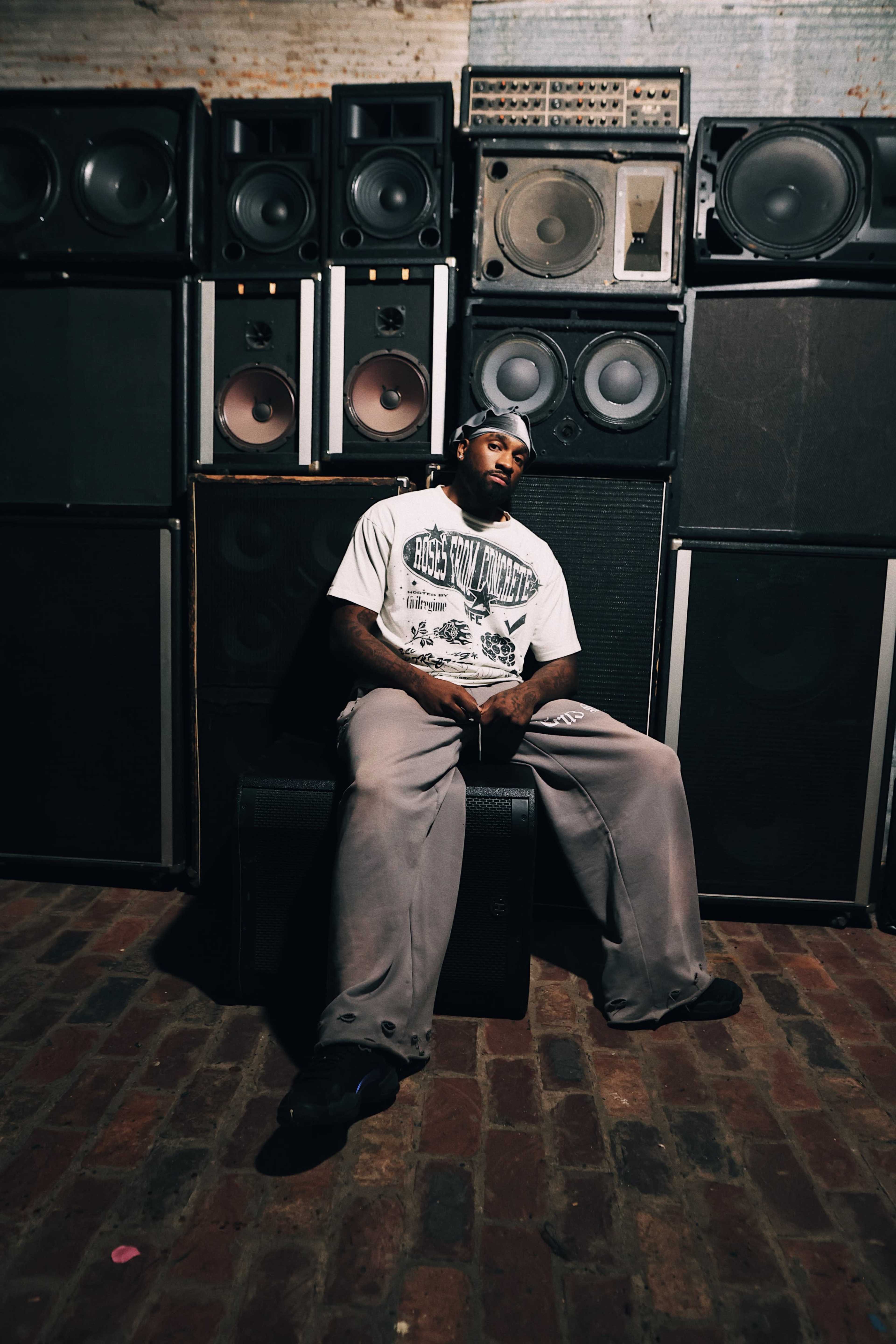 A man sits on a crate in front of a wall of amplifiers and speakers in a dimly lit room.