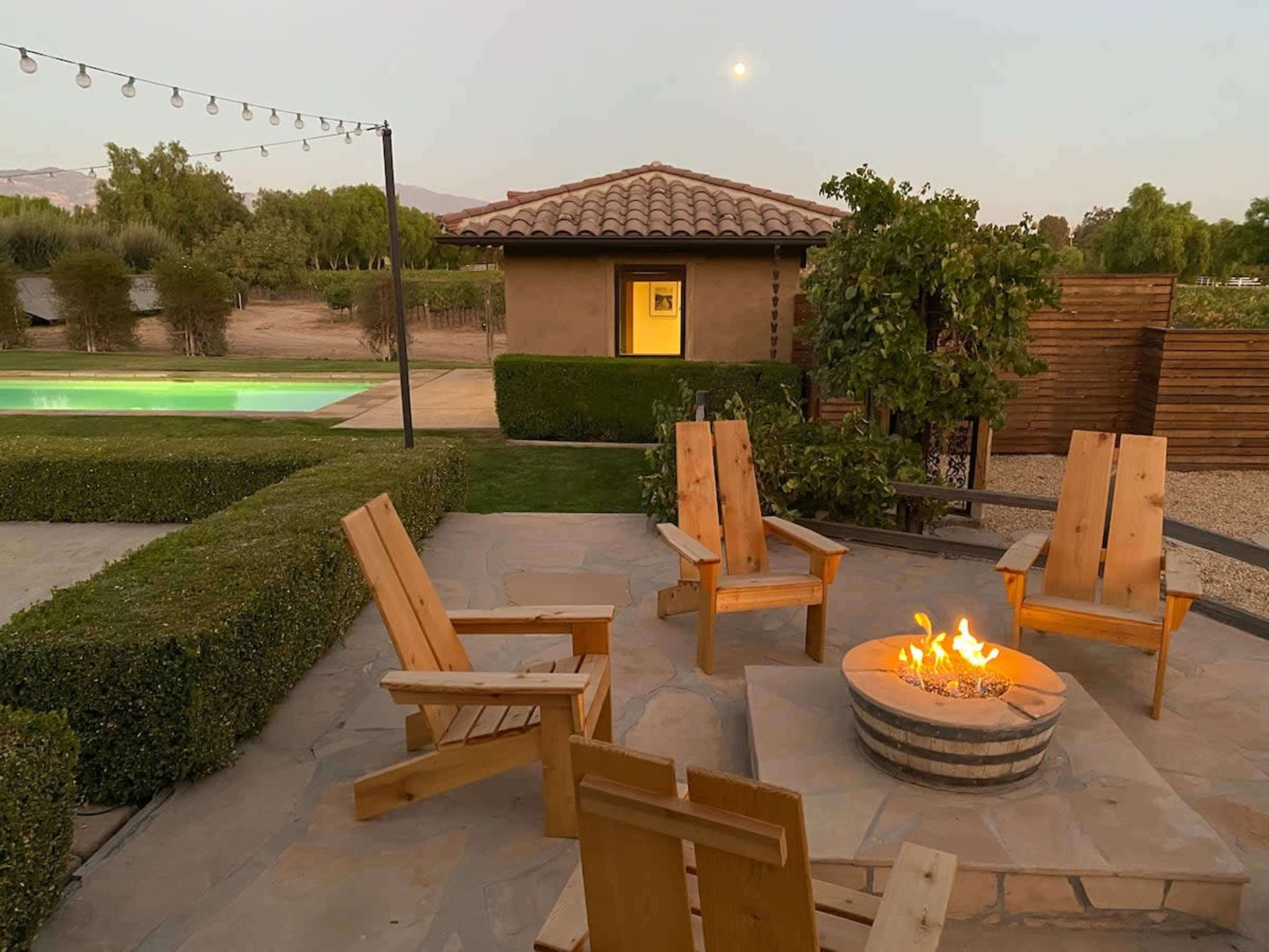 A fire pit surrounded by wooden chairs is situated in a backyard with a pool and a house in the background.