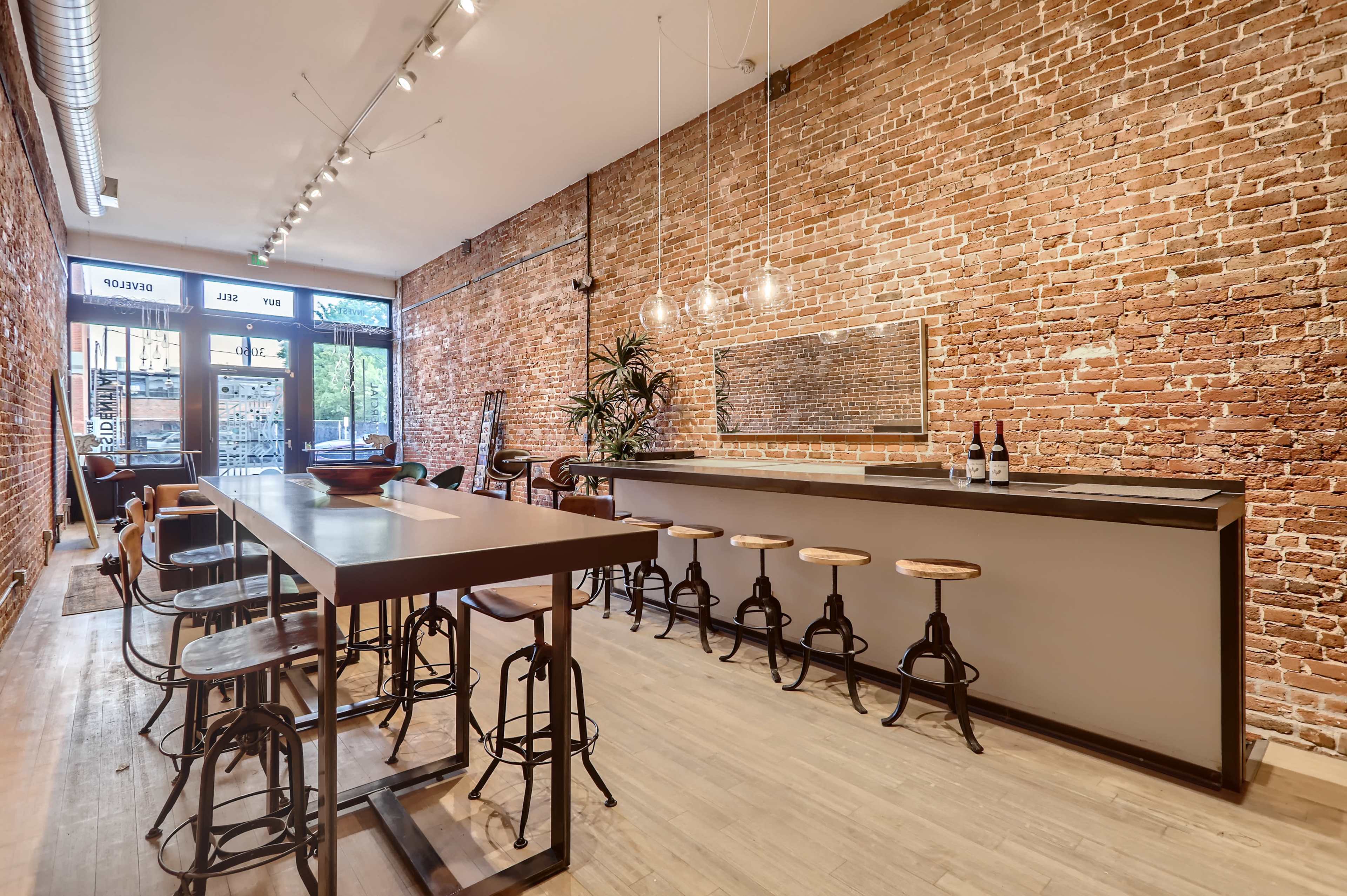 A modern interior of a restaurant or bar with exposed brick walls, a long table in the center, and barstools arranged along a counter.