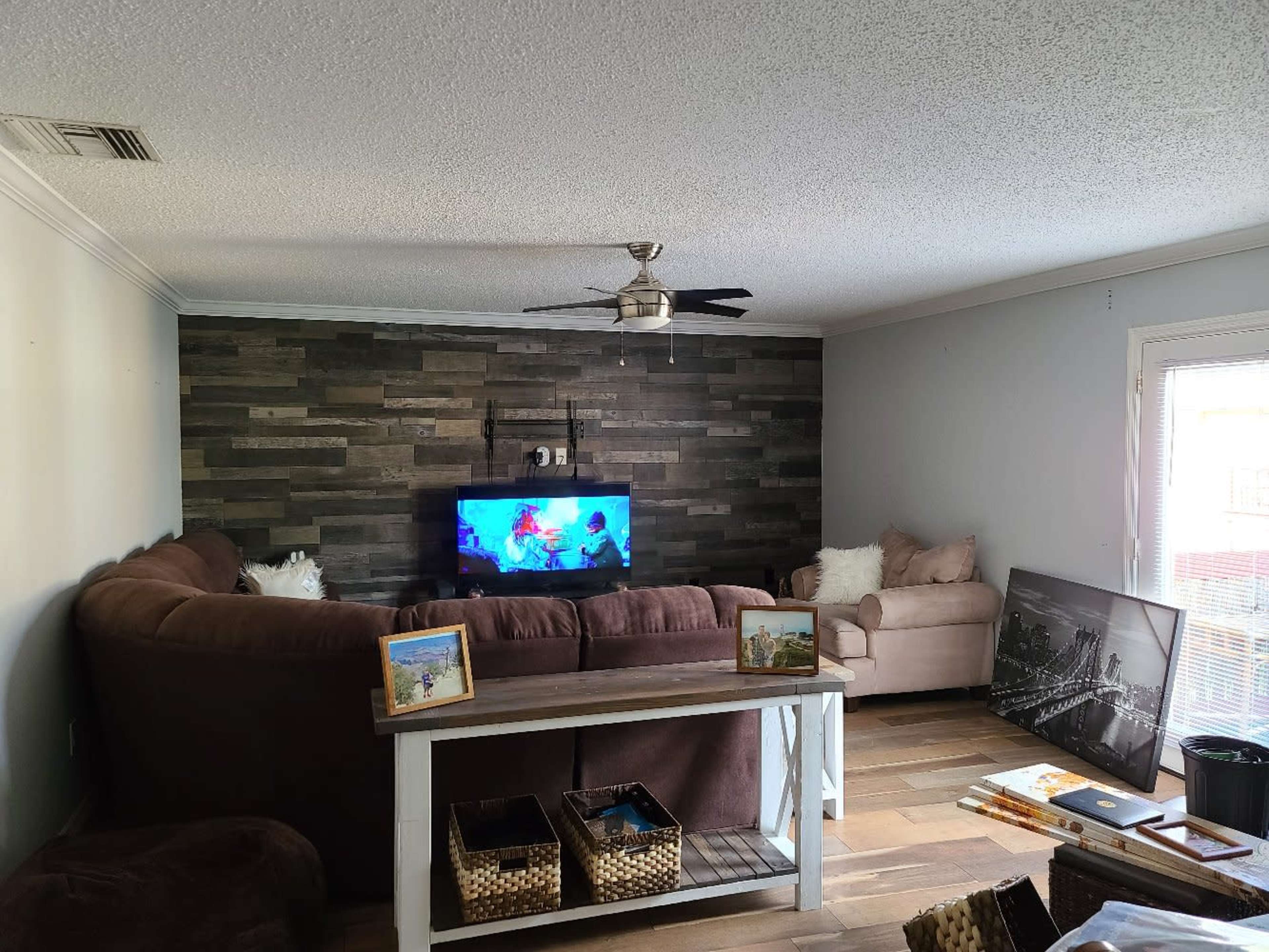The living room features a large sectional couch facing a television mounted against a textured wall, with a coffee table in front and a casual seating area to the side.