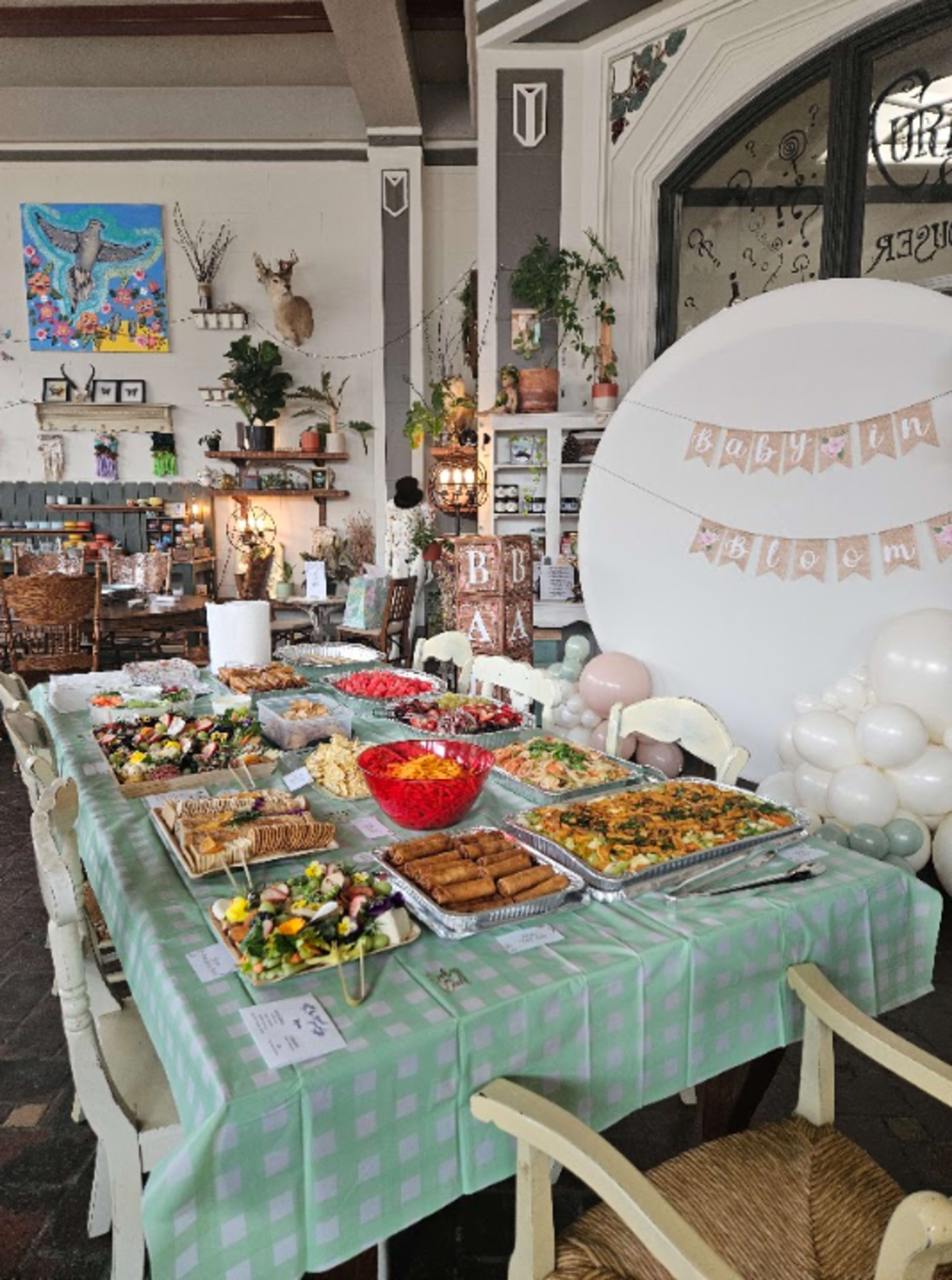 The image shows a decorated table set for a celebration, featuring a variety of dishes, including salads, pasta, and pastries, arranged under a banner that reads "Baby in Bloom."