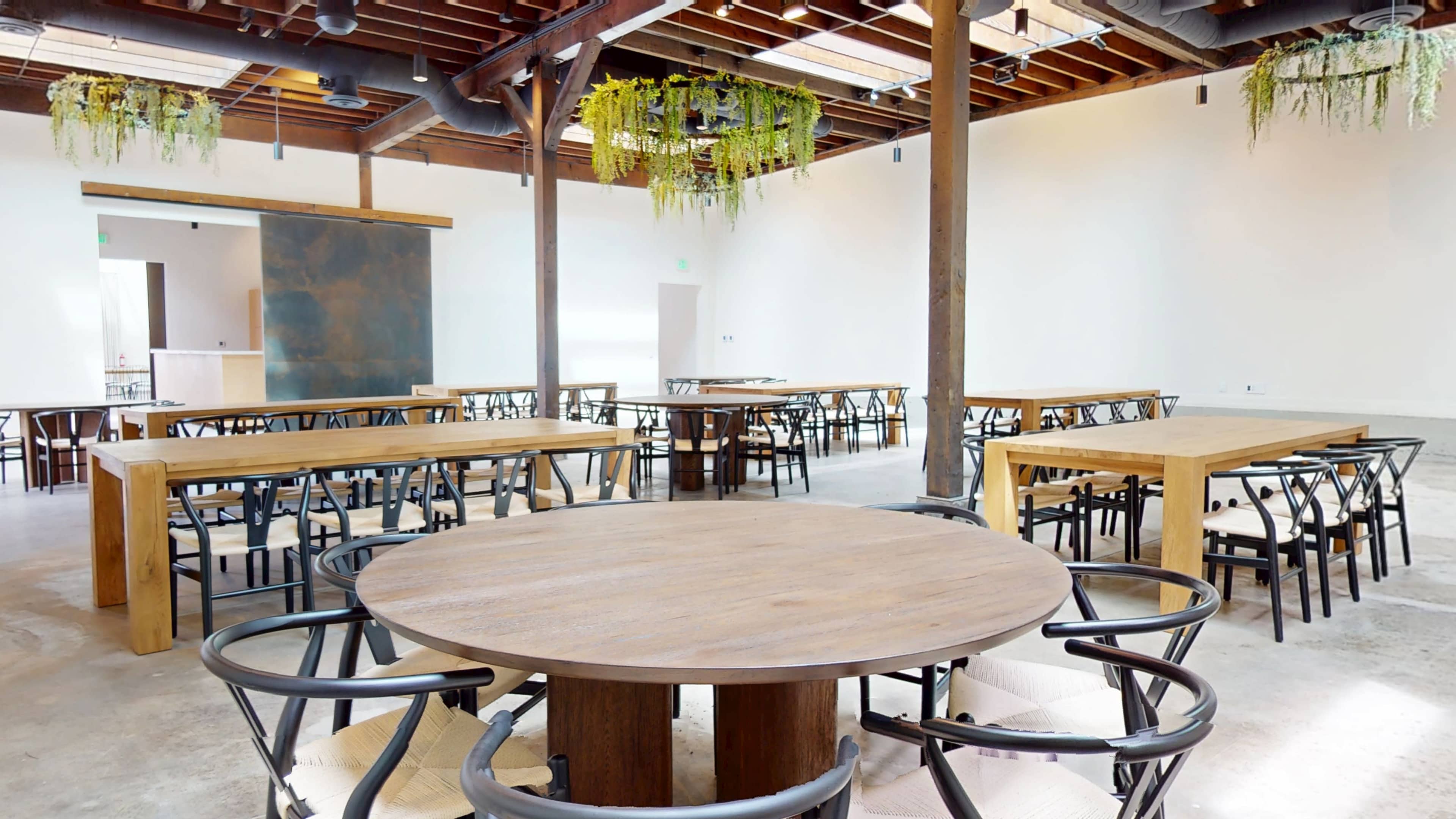 The image shows a spacious dining area featuring wooden tables and chairs with exposed wooden beams and hanging plants overhead.