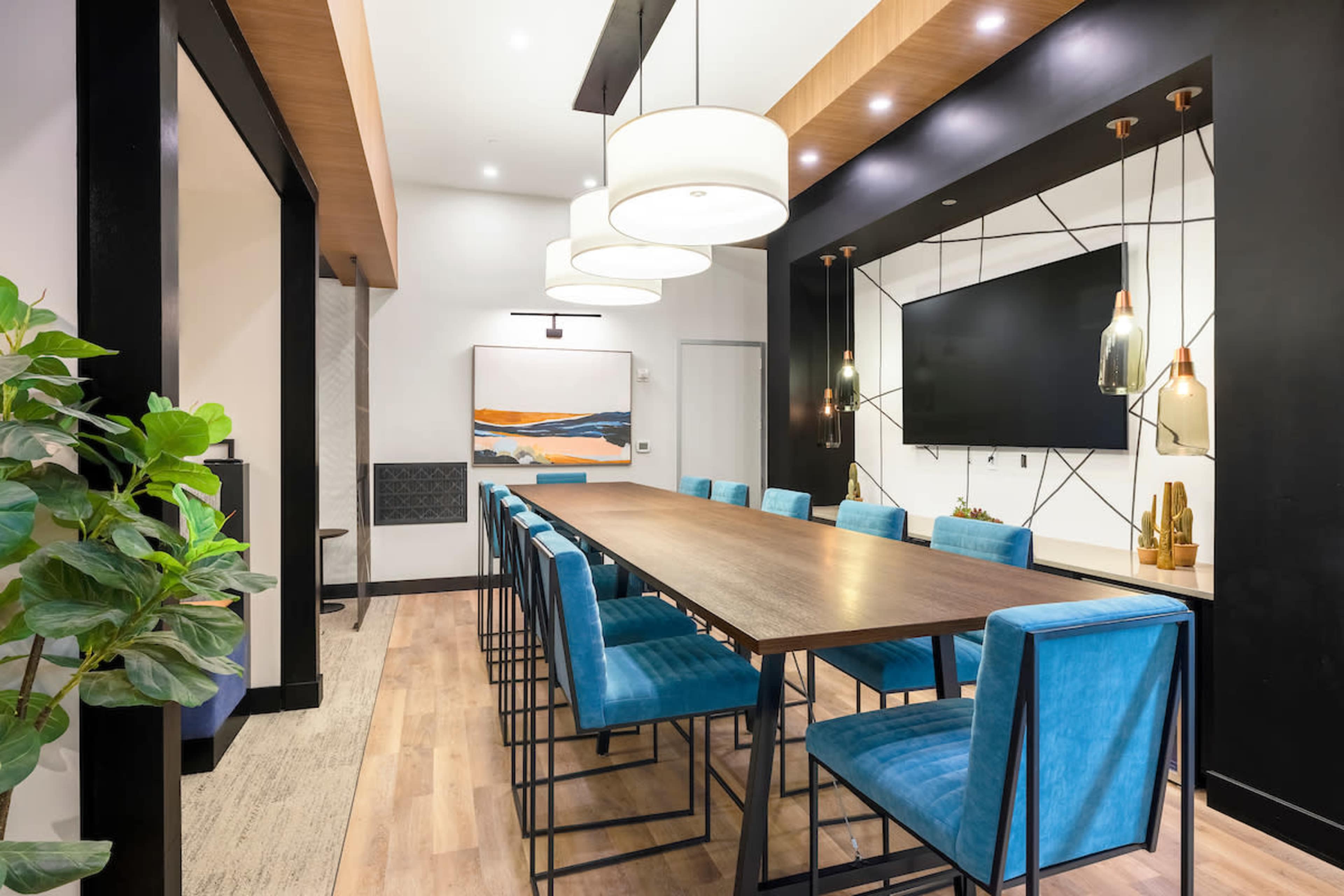 A long wooden table is surrounded by blue upholstered chairs in a well-lit dining area featuring a television and decorative wall art.