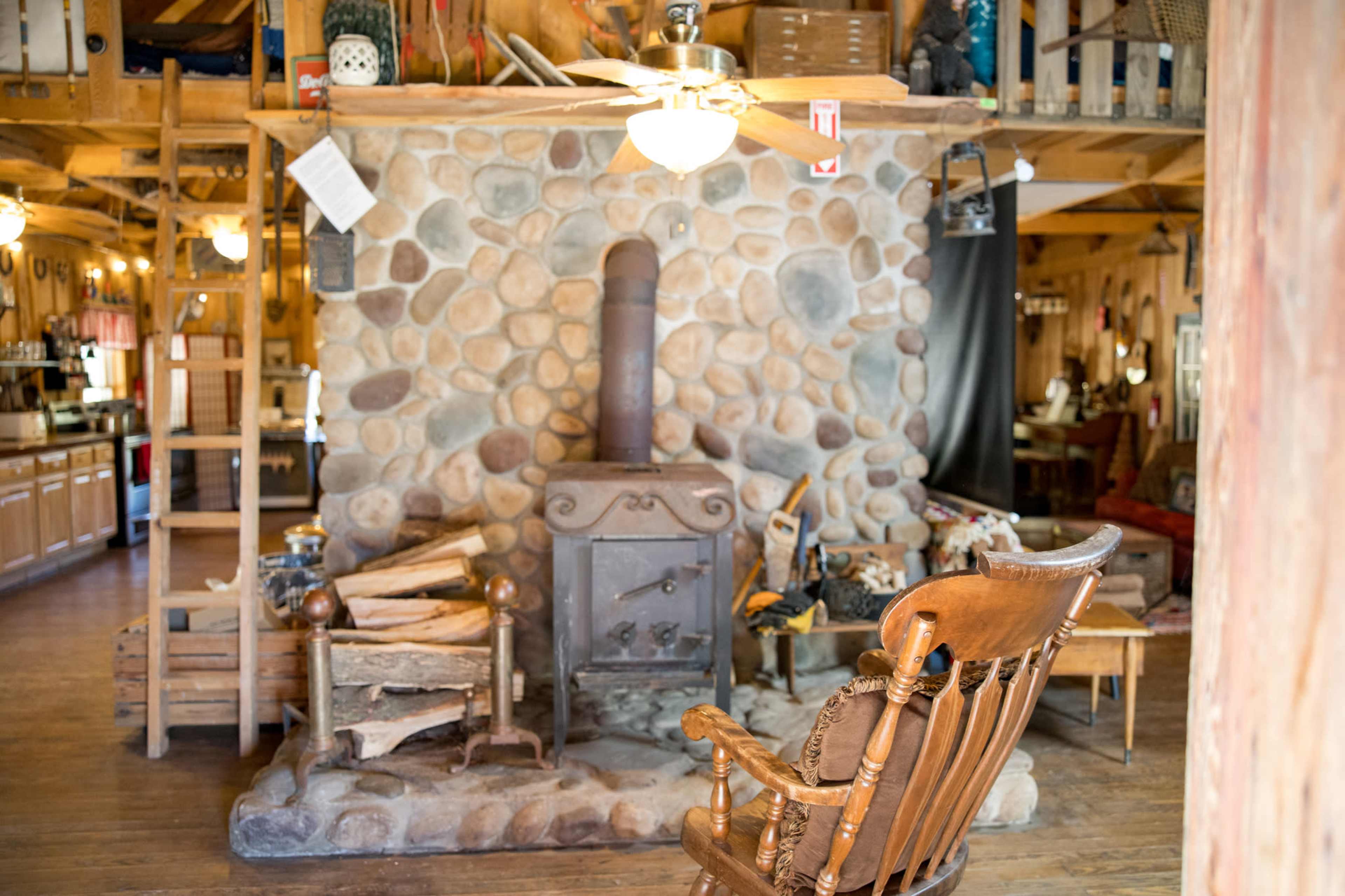 The image shows a rustic interior featuring a stone fireplace, a wooden rocking chair, and various decorations in a cozy cabin.