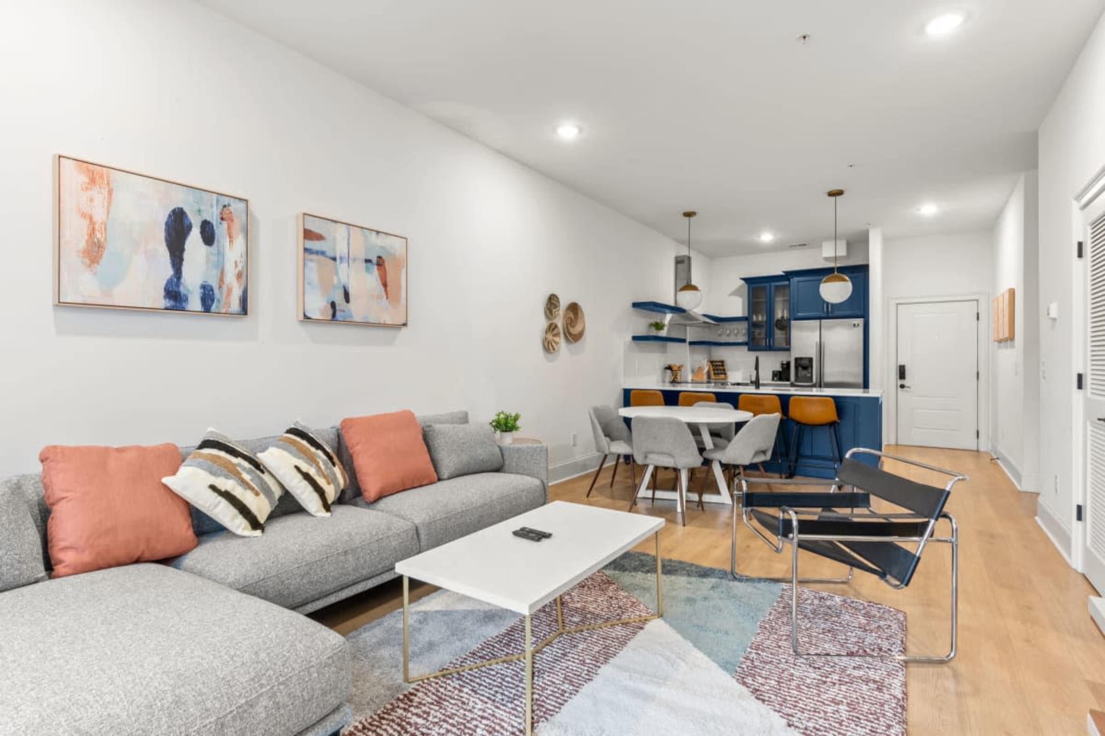 The living area features a gray sectional sofa with colorful pillows, a white coffee table, and access to a modern kitchen with blue cabinets and bar seating.