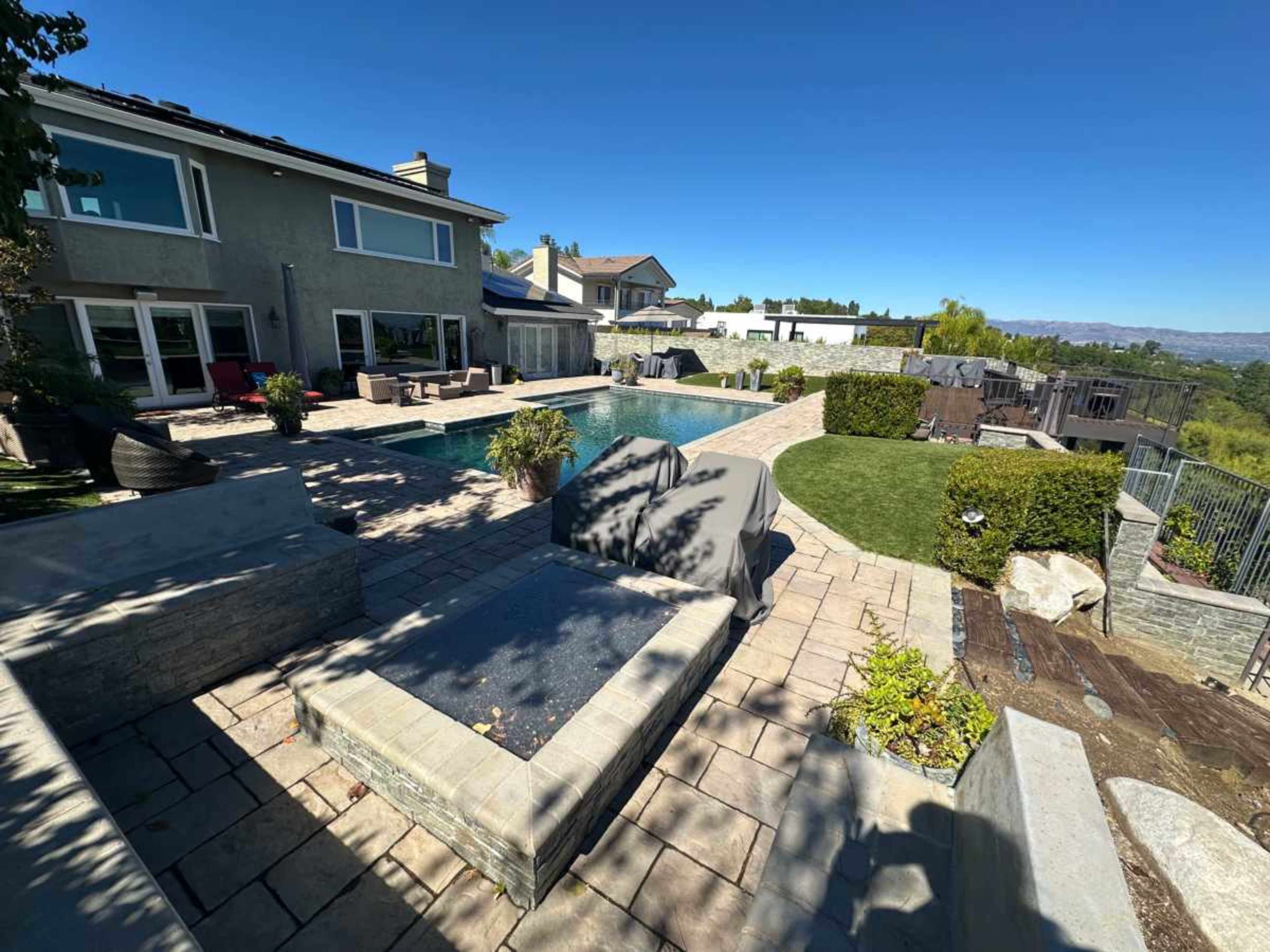 The image shows a spacious backyard with a swimming pool, a patio area, and landscaped greenery surrounding a barbecue grill.