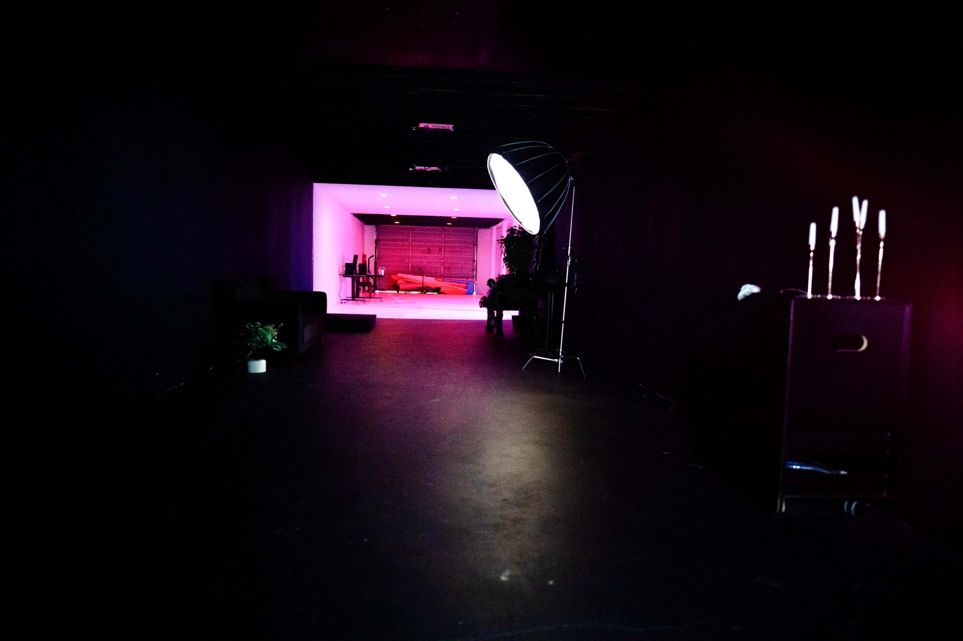 A dimly lit hallway leads to a brightly lit room with purple walls and a red sofa, featuring a photography light on the side.