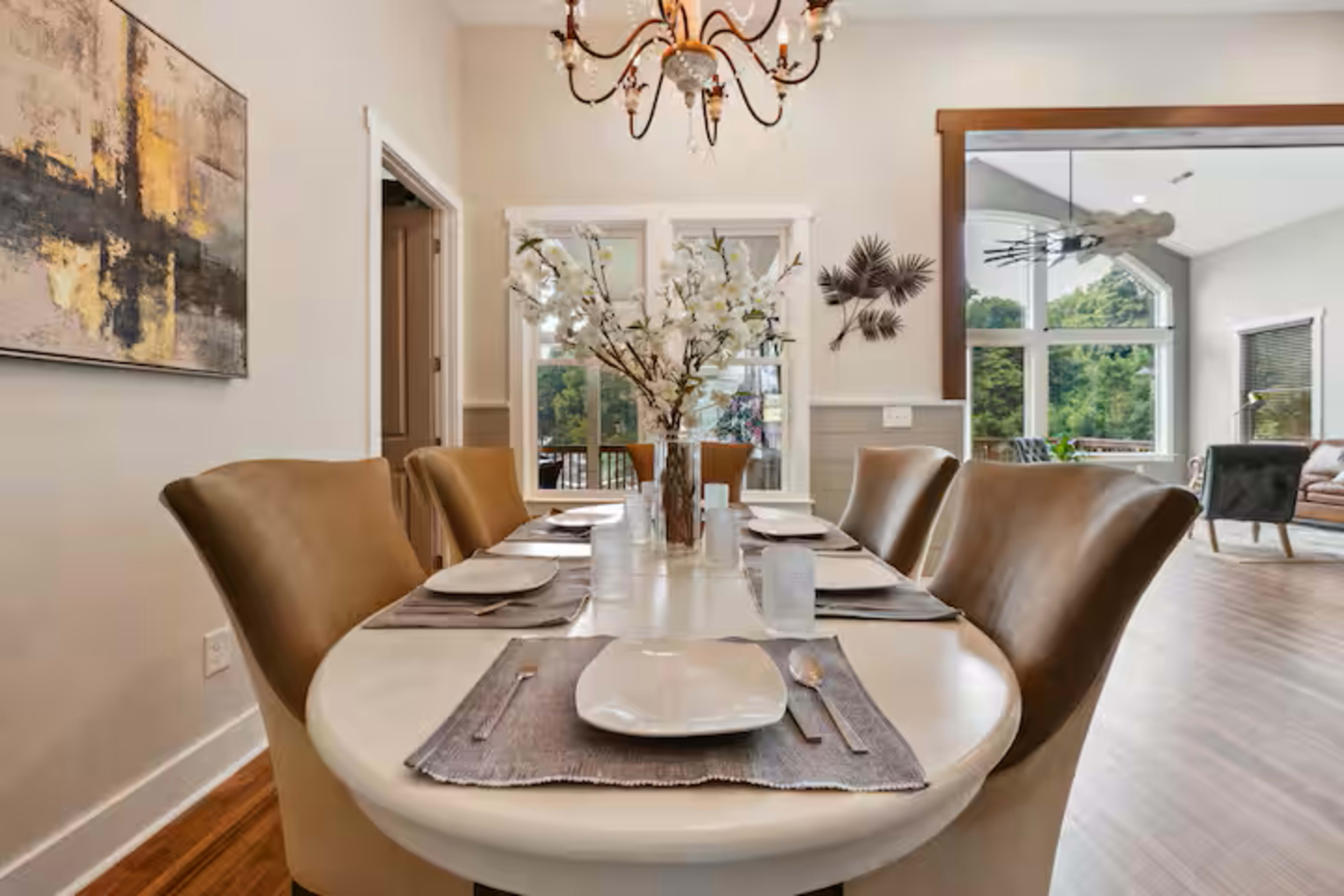 A dining table is set with plates and glasses, surrounded by upholstered chairs, beneath a chandelier in a room with large windows overlooking greenery.