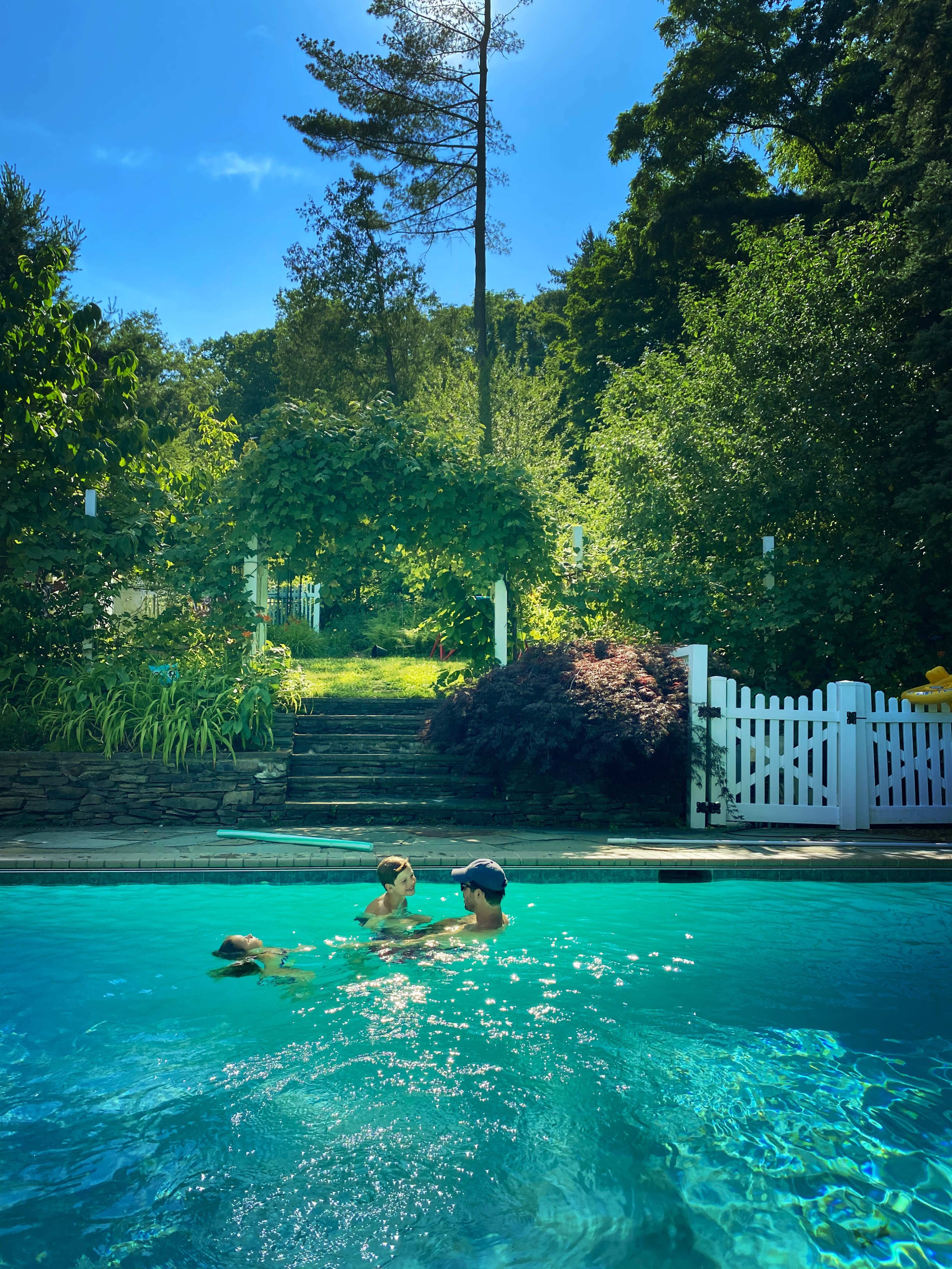A man and a child swim in a blue pool surrounded by lush greenery and a white picket fence.
