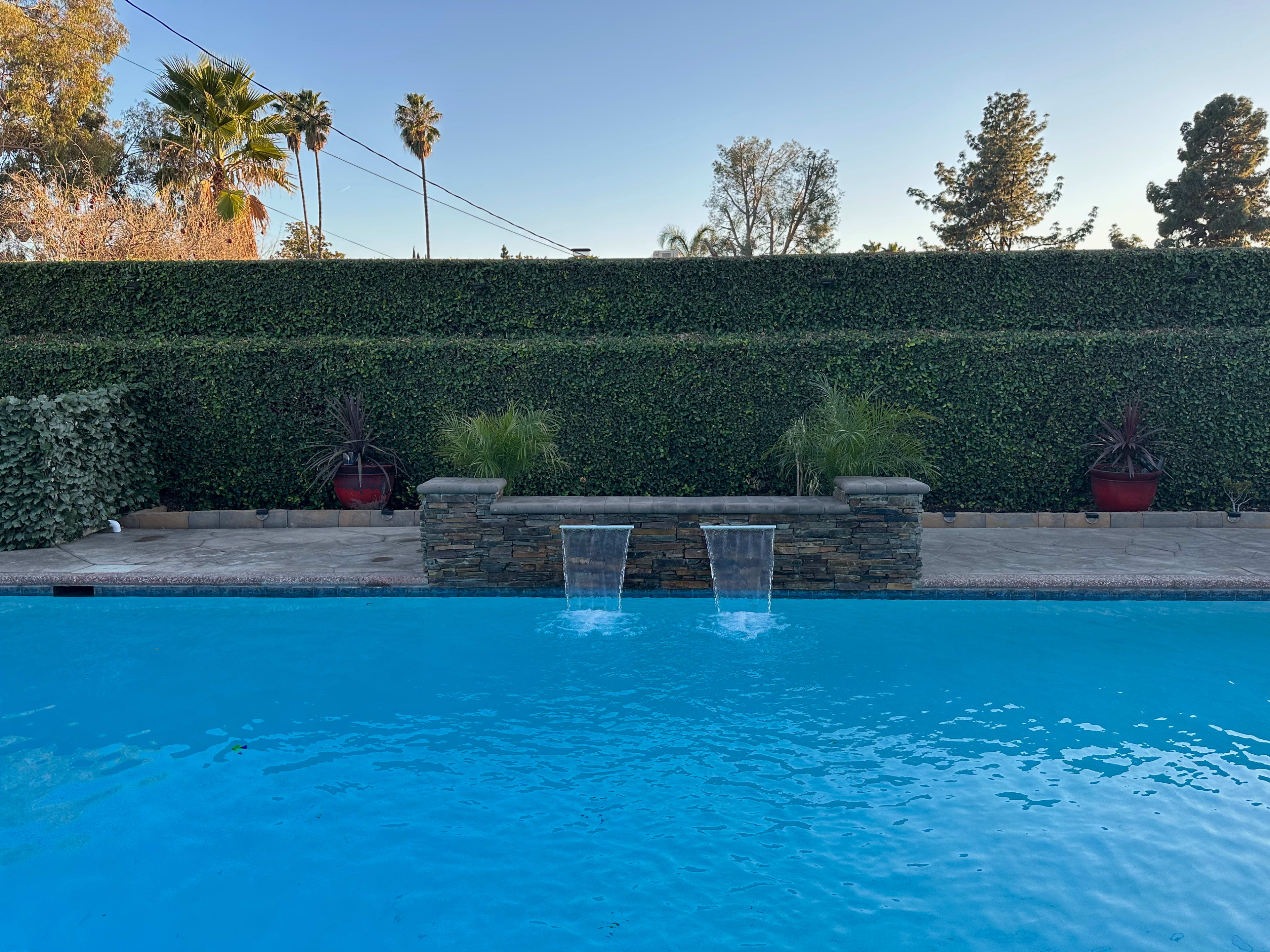 A swimming pool with two water features spilling into it, surrounded by a neatly trimmed hedge and decorative plants in pots.