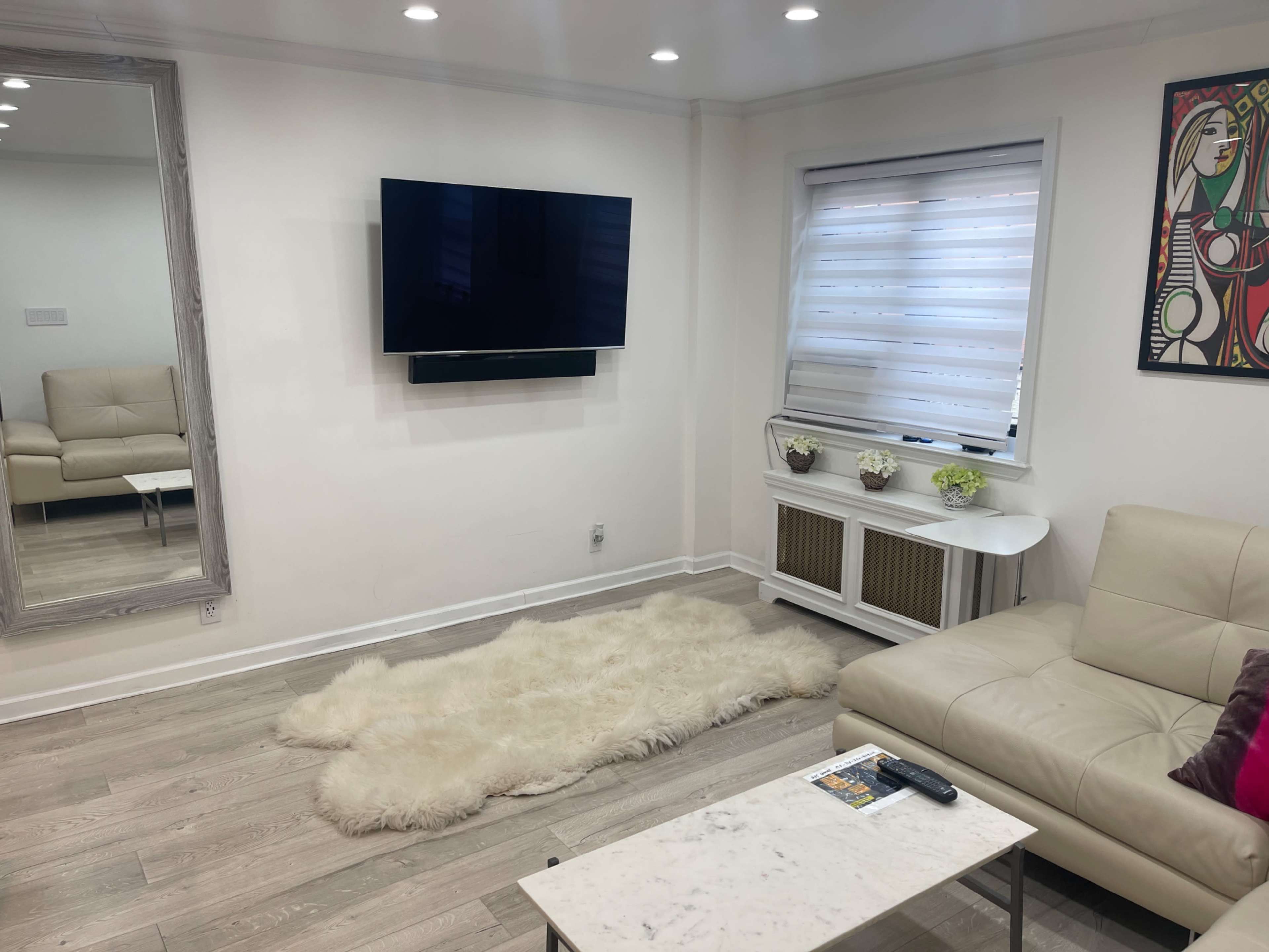 A living room featuring a beige leather sofa, a wall-mounted television, a white coffee table, and a large mirror, all set against light-colored walls and wooden flooring.