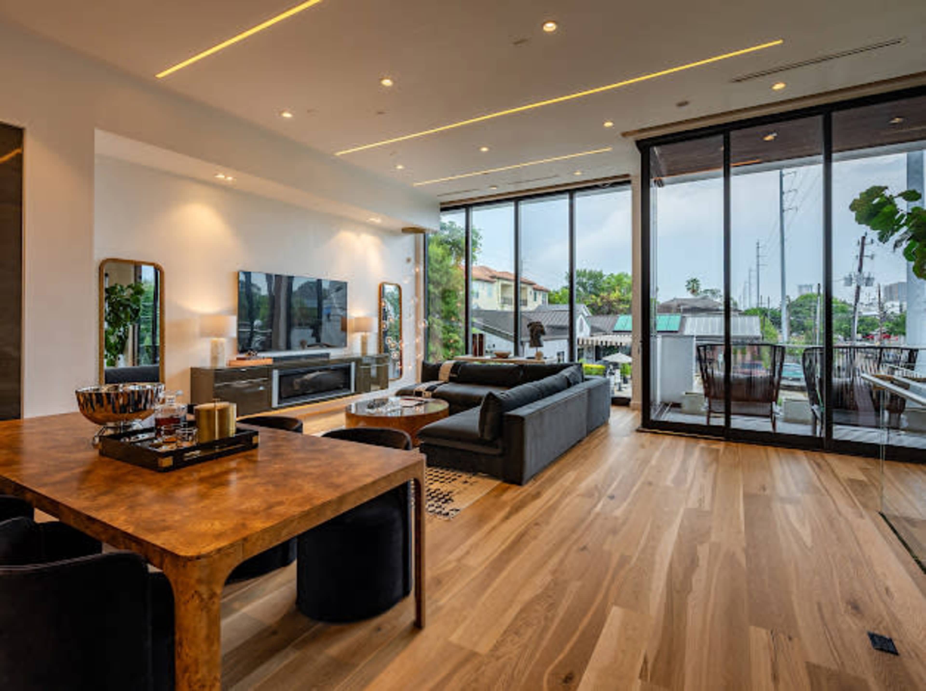The image shows a modern living room with large windows, a dining area with a wooden table, a black sofa, and a television mounted on the wall.
