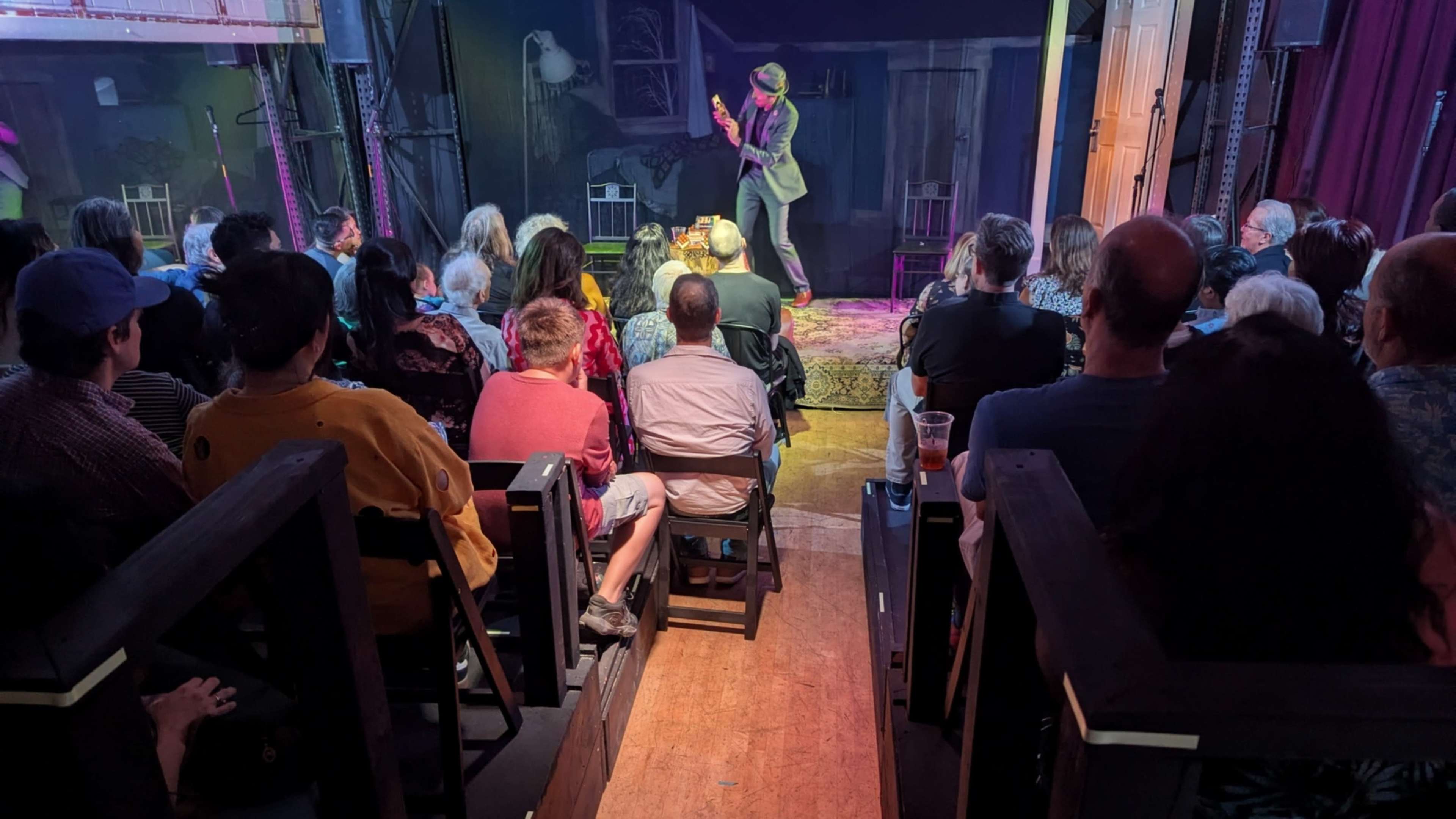 A performer in a suit plays guitar on stage while an audience watches from seated rows in a dimly lit venue.