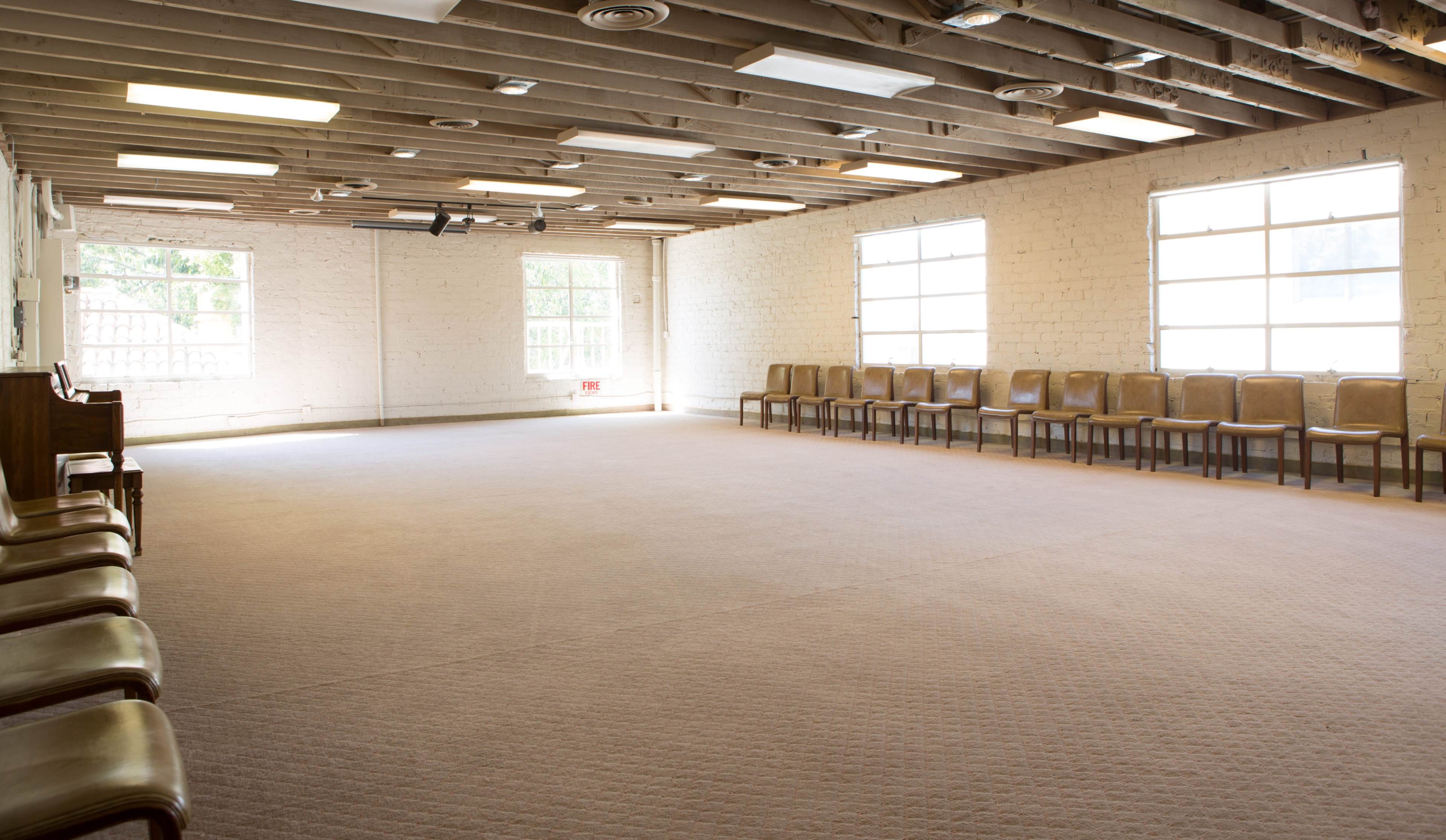 An empty room with wooden beams, carpeted flooring, and a row of wooden chairs along one wall.