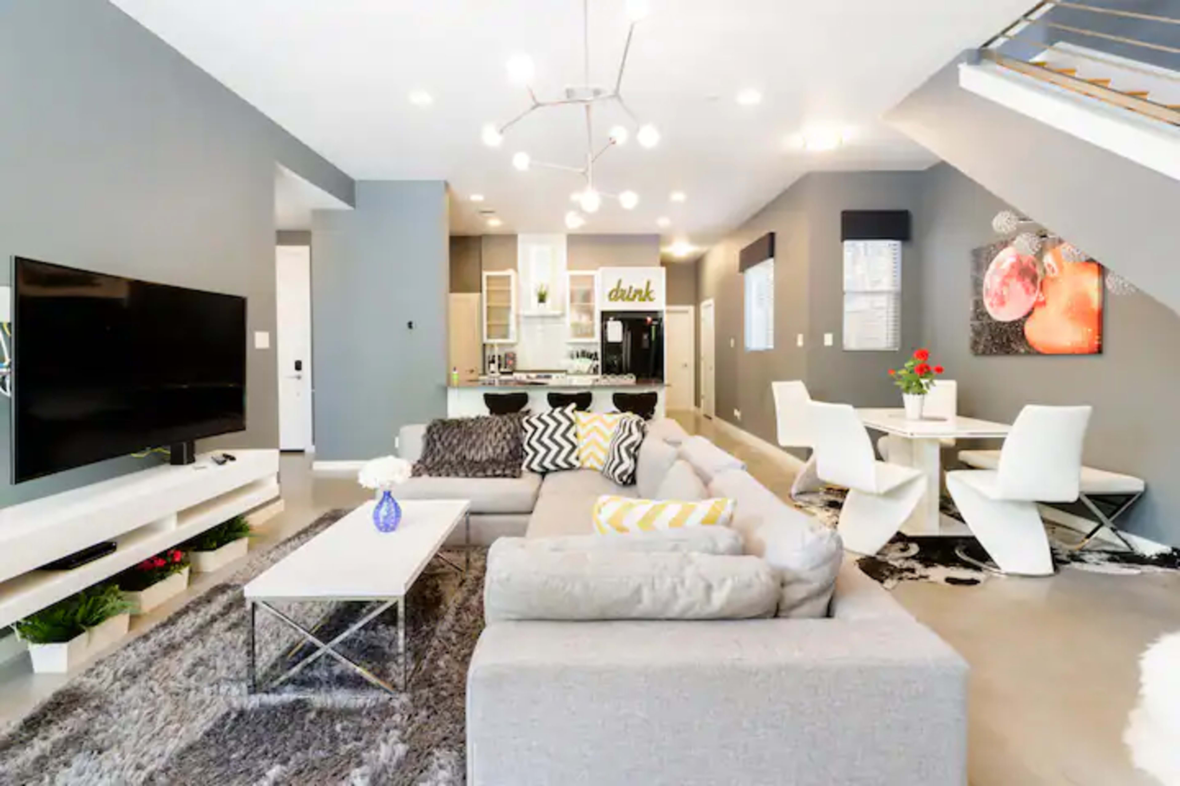 The image shows a modern living room with a gray sectional sofa, a coffee table, and a dining area featuring white chairs and a table, all illuminated by overhead lights.