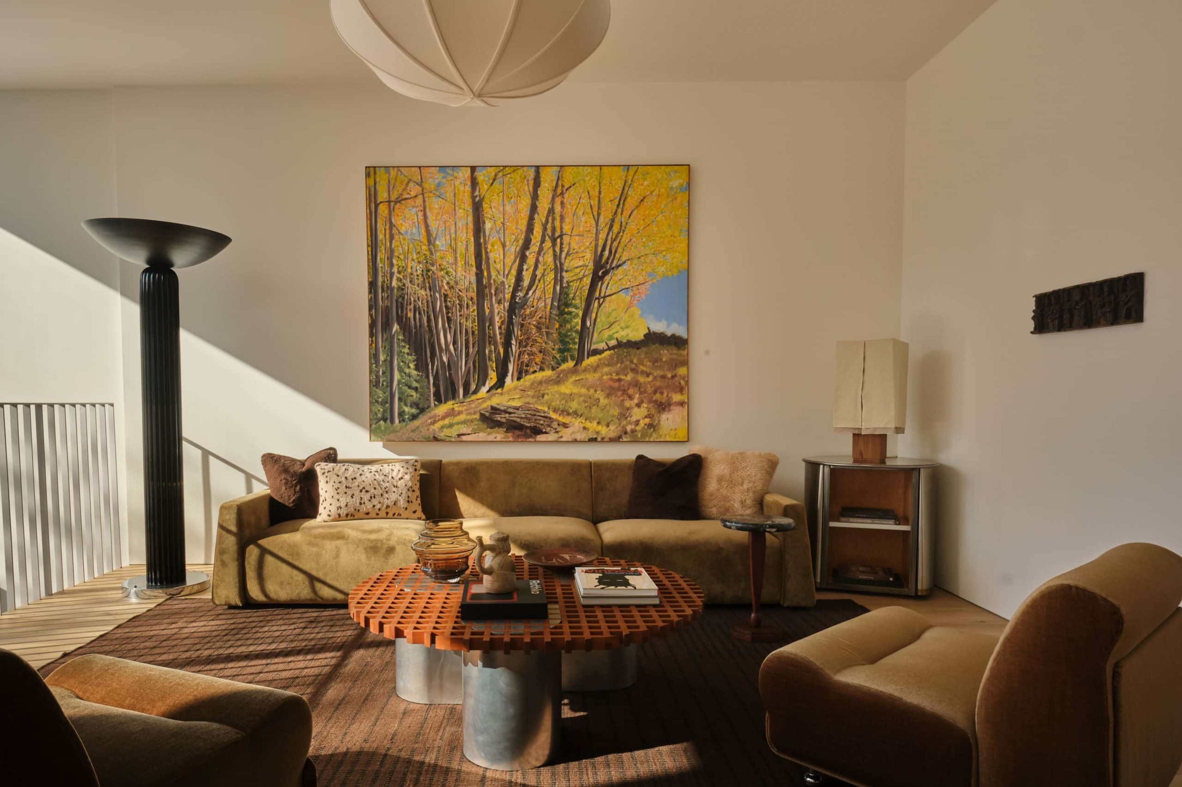 The image shows a living room featuring a large painting of trees on the wall, a circular coffee table in the center, and various seating options arranged around it.