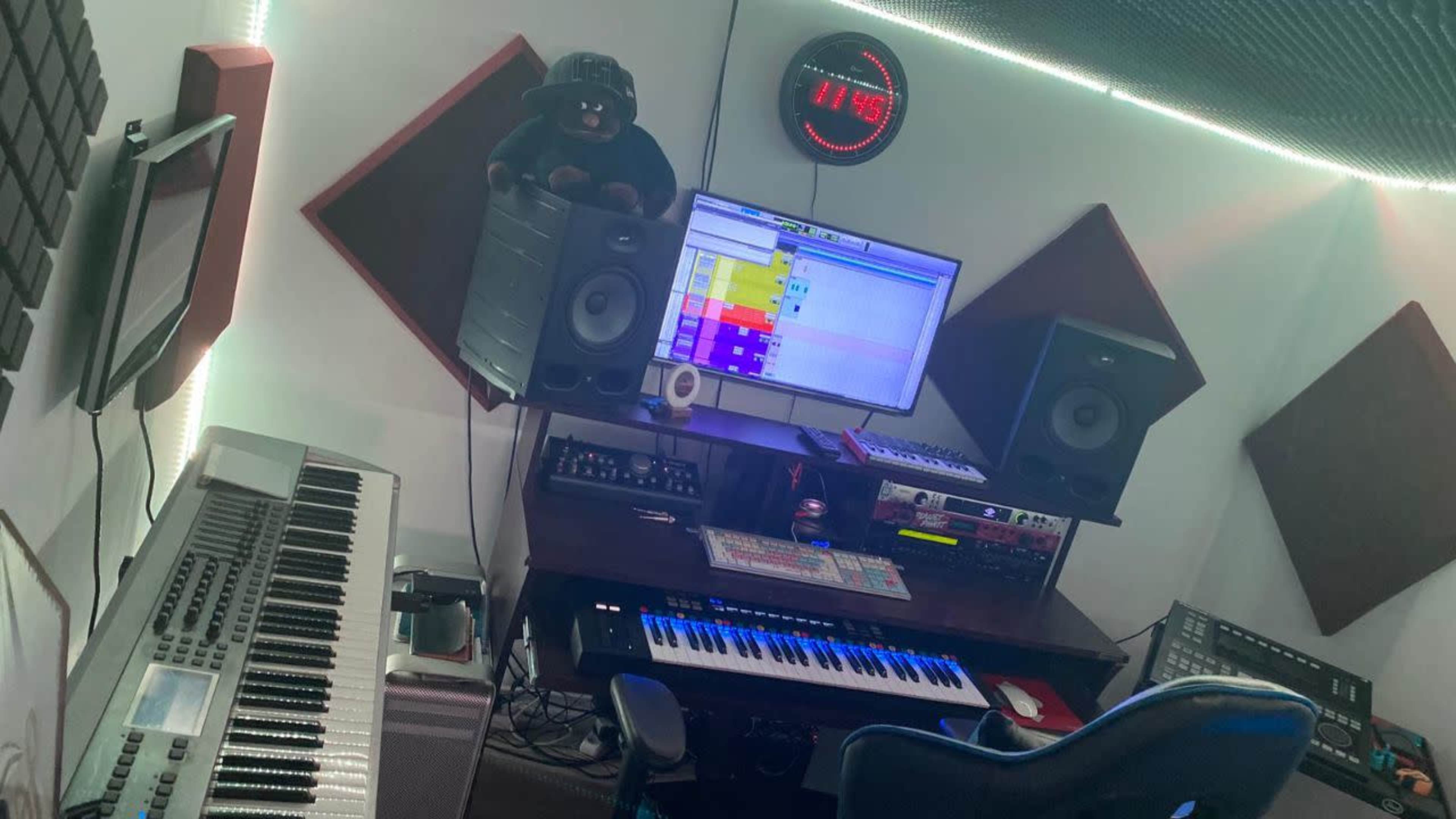 A music production workspace featuring a keyboard, speakers, a computer monitor with software open, and acoustic panels on the walls.