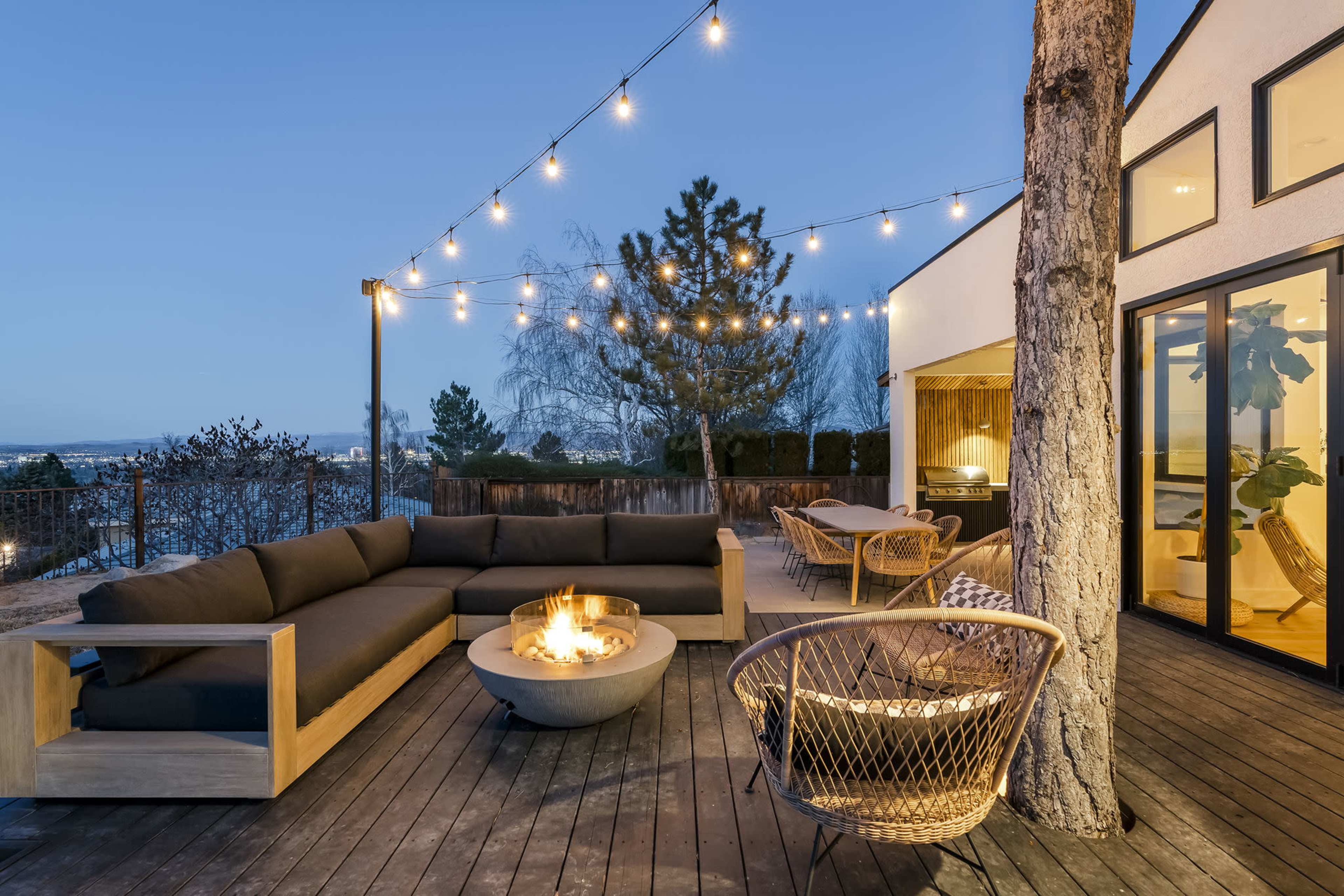 A modern outdoor living space features a sectional sofa surrounding a fire pit, illuminated by string lights at dusk.