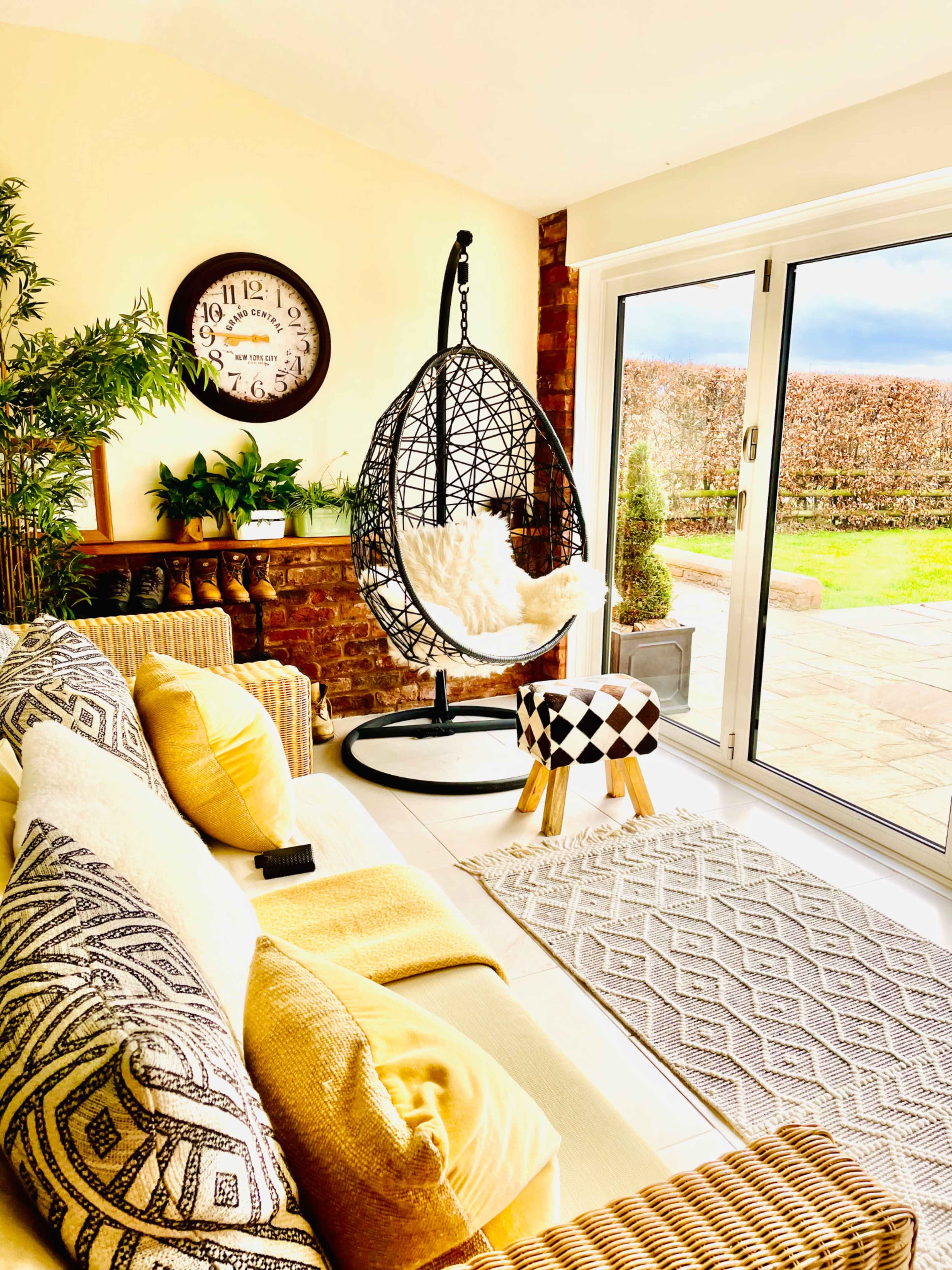 The image depicts a cozy living room with a black hanging chair, a patterned stool, and large windows overlooking a garden.