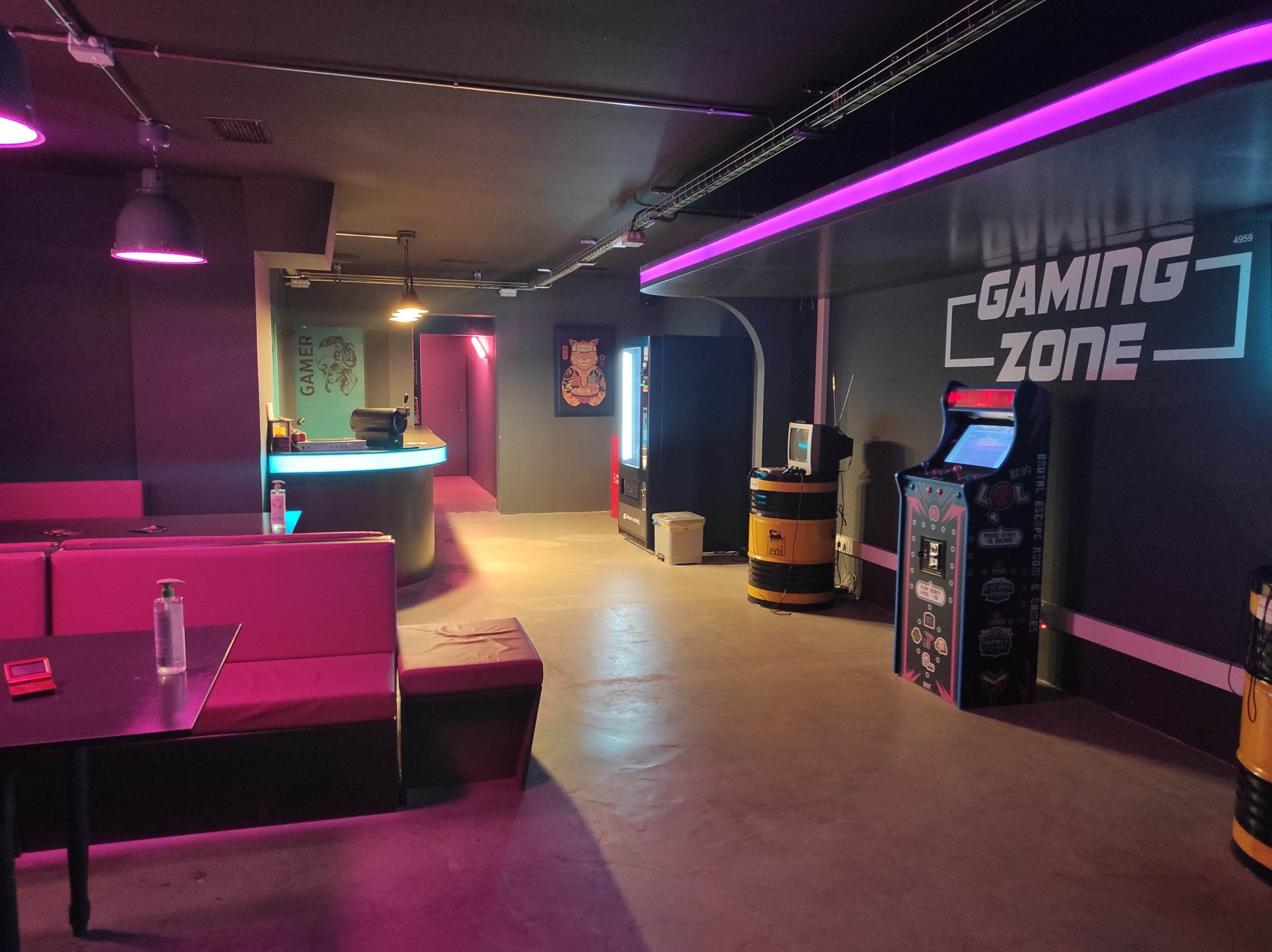 A dimly lit gaming zone with neon lights, featuring seating areas, vending machines, and arcade cabinets.