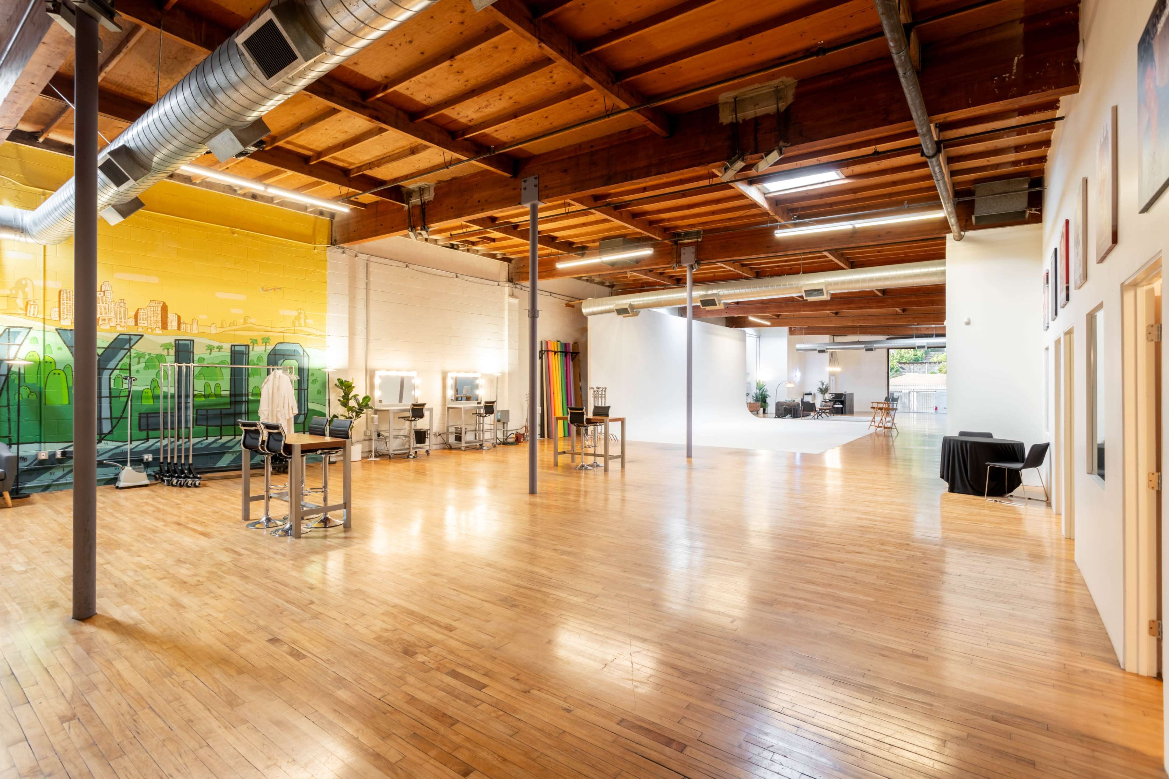 The image depicts a spacious studio with wooden floors, high ceilings, and various workstations, including mirrors and lighting equipment, against a colorful mural.