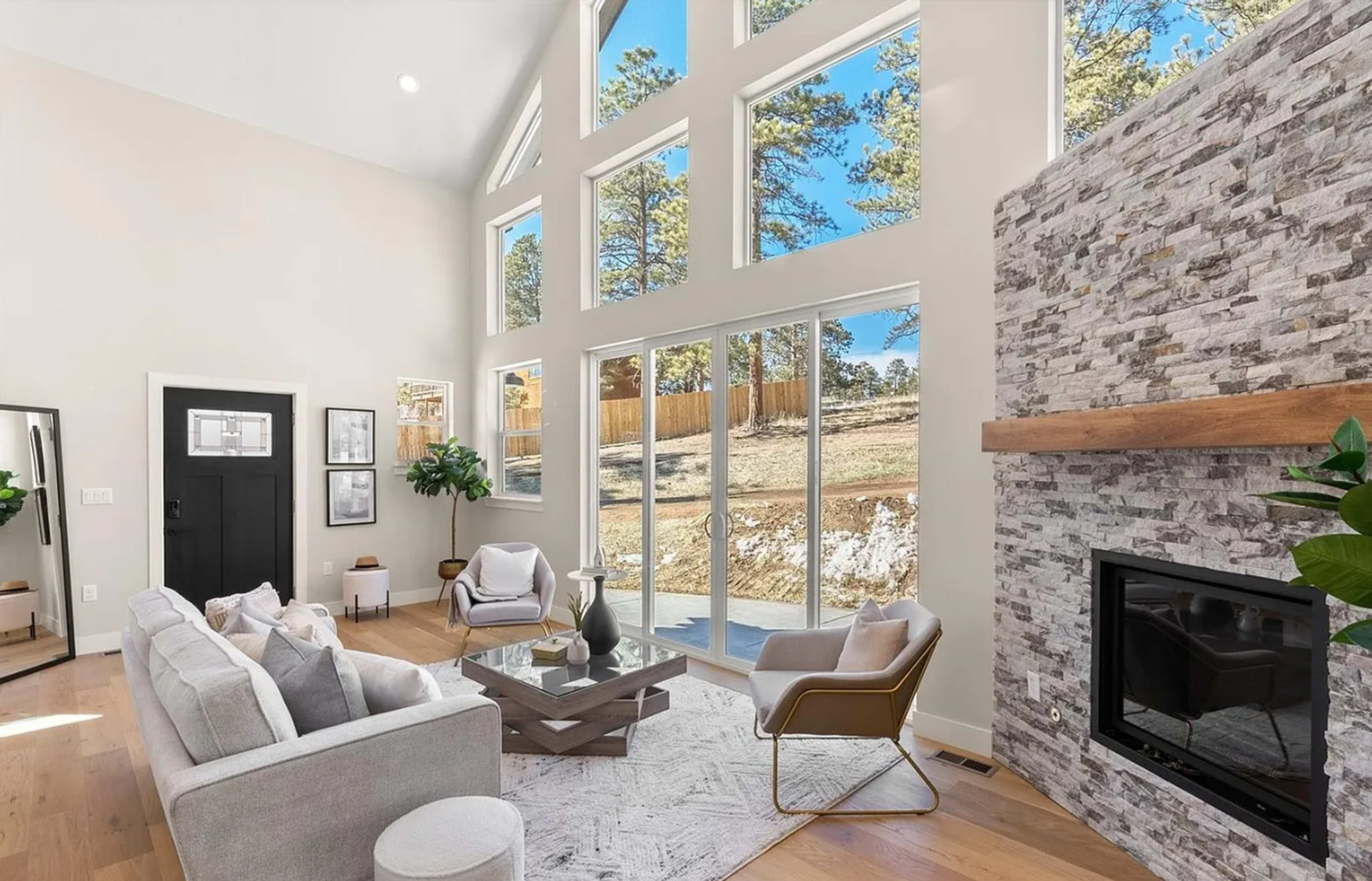 The image shows a modern living room with large windows, a stone fireplace, and contemporary furniture, overlooking a wooded area.