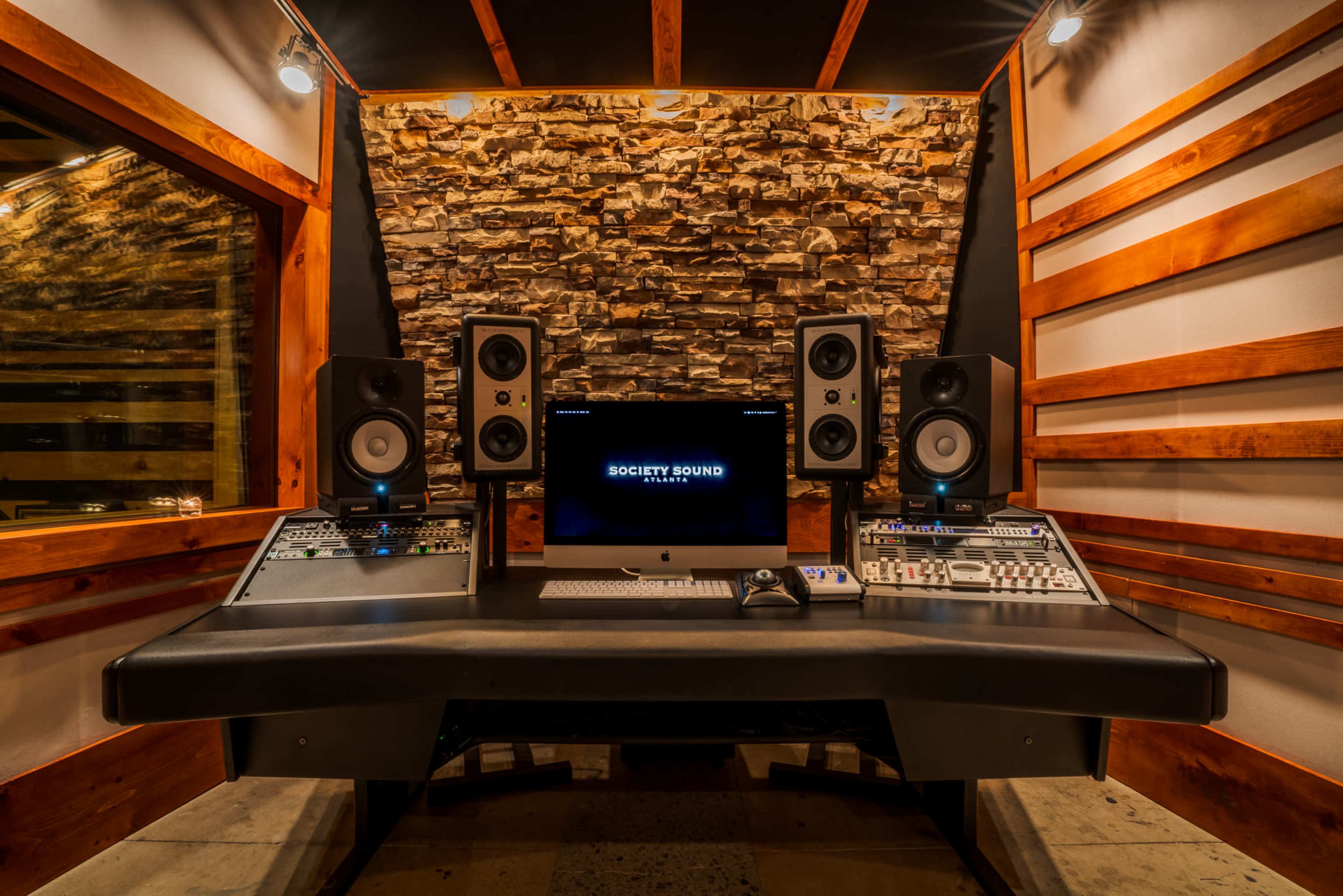 The image shows a recording studio with a control desk, speakers, and a computer screen displaying "SOCIETY SOUND" against a stone wall backdrop.