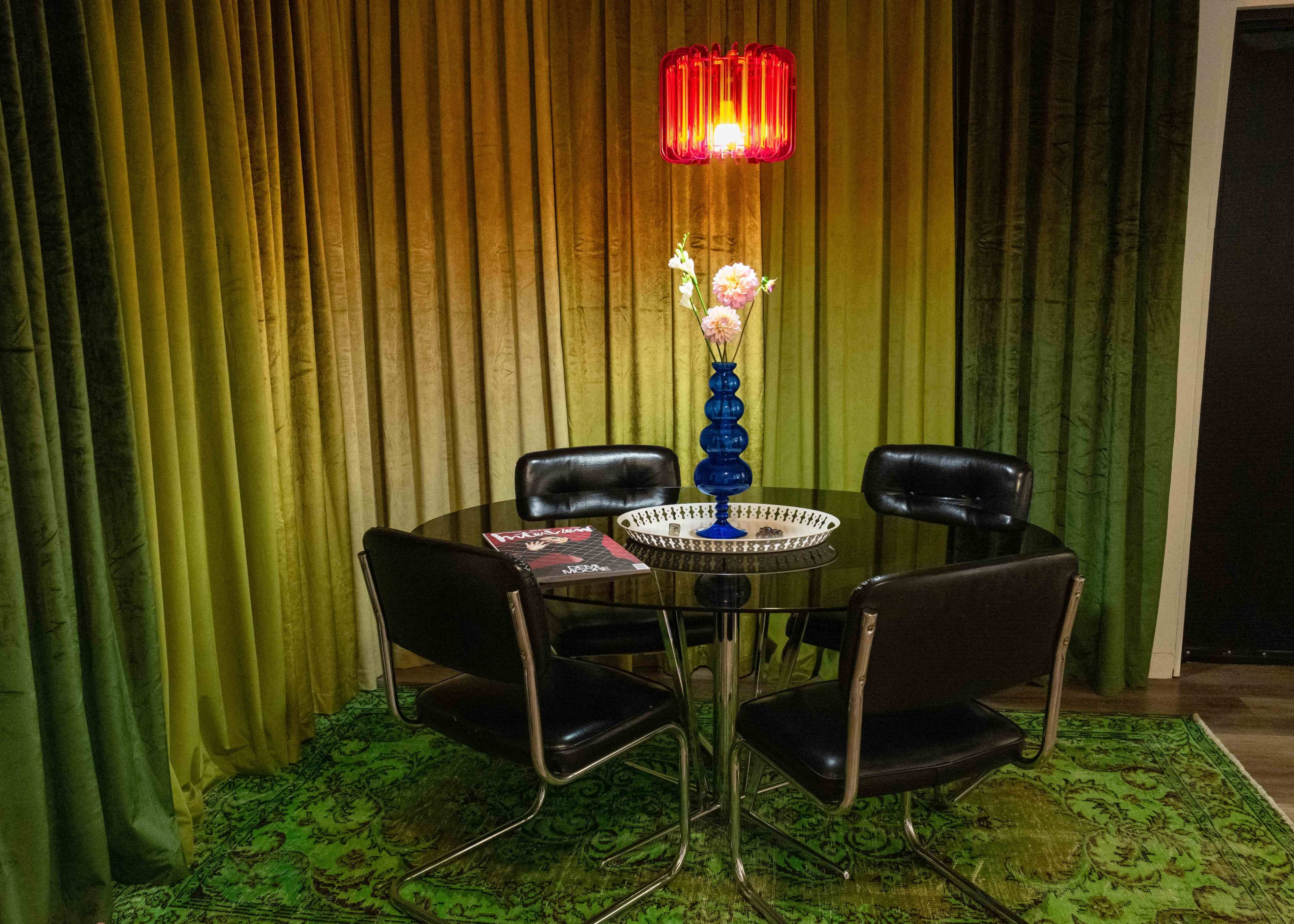 A round black table with three black chairs is set against a backdrop of green curtains, illuminated by a red pendant light, and features a blue vase with flowers on the table.