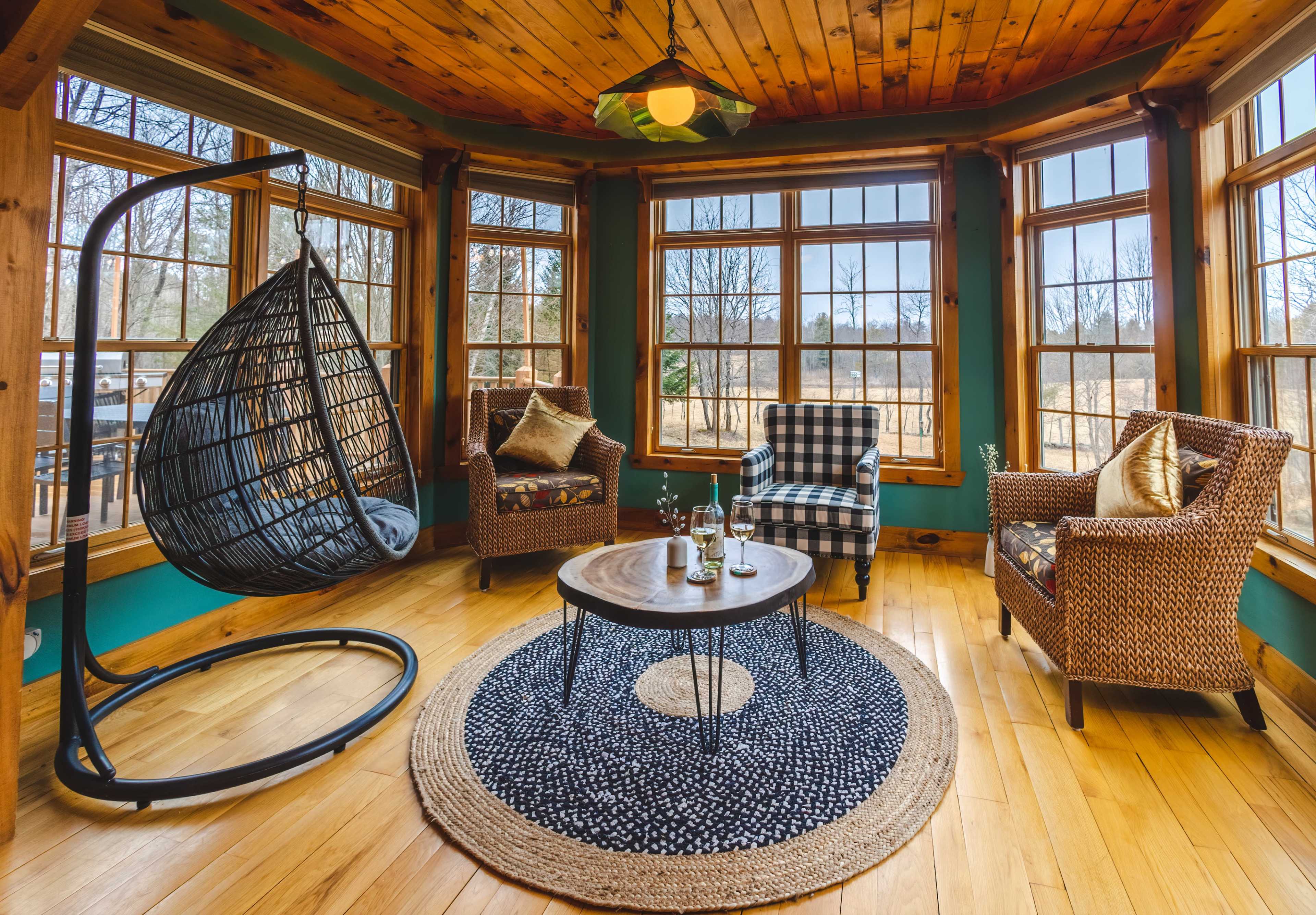 The scene features a cozy sunroom with a hanging chair, two patterned armchairs, a round table, and large windows allowing natural light to fill the space.
