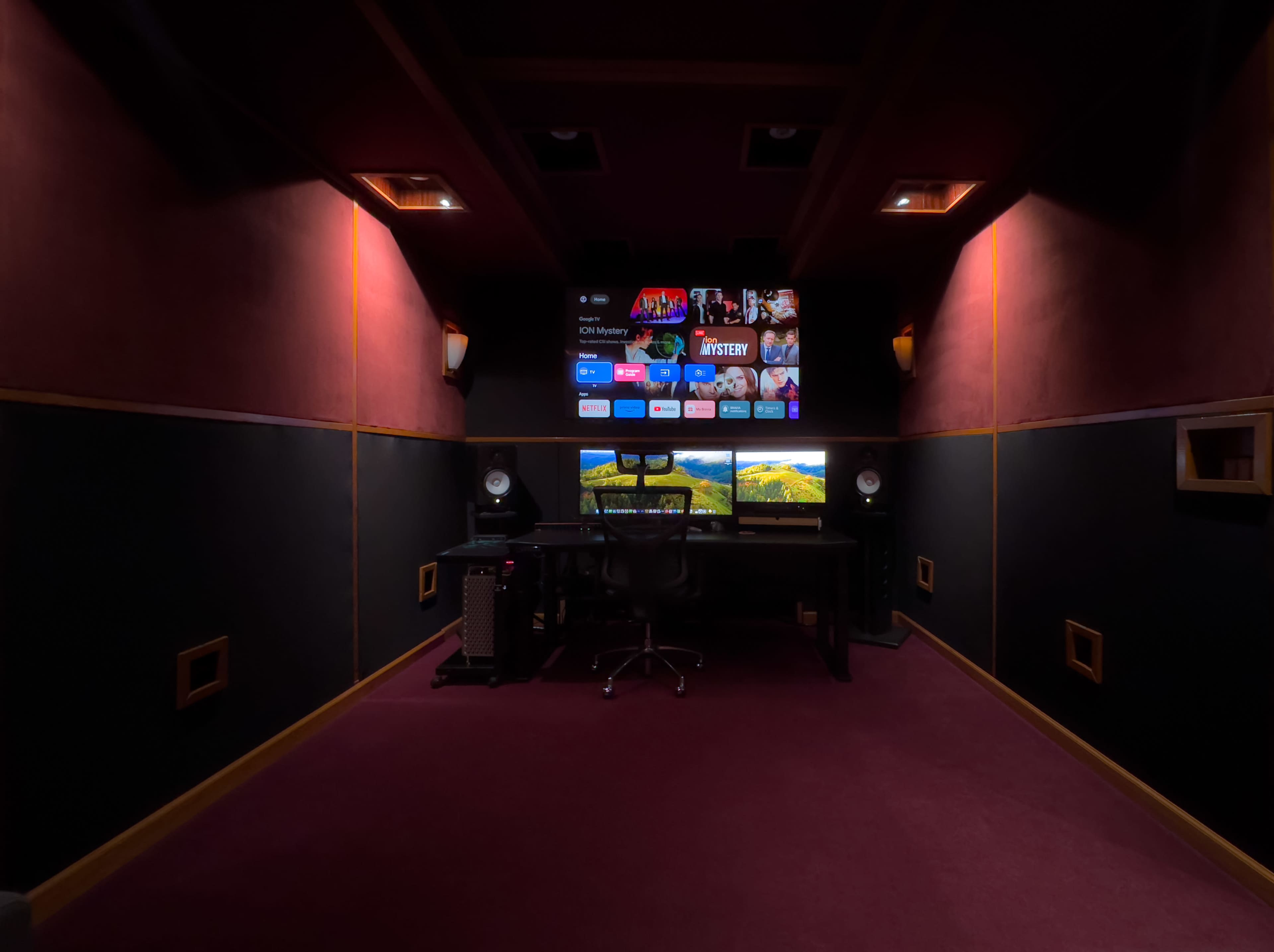 A dimly lit media room with a large screen displaying streaming options and a desk setup featuring two monitors and audio speakers.