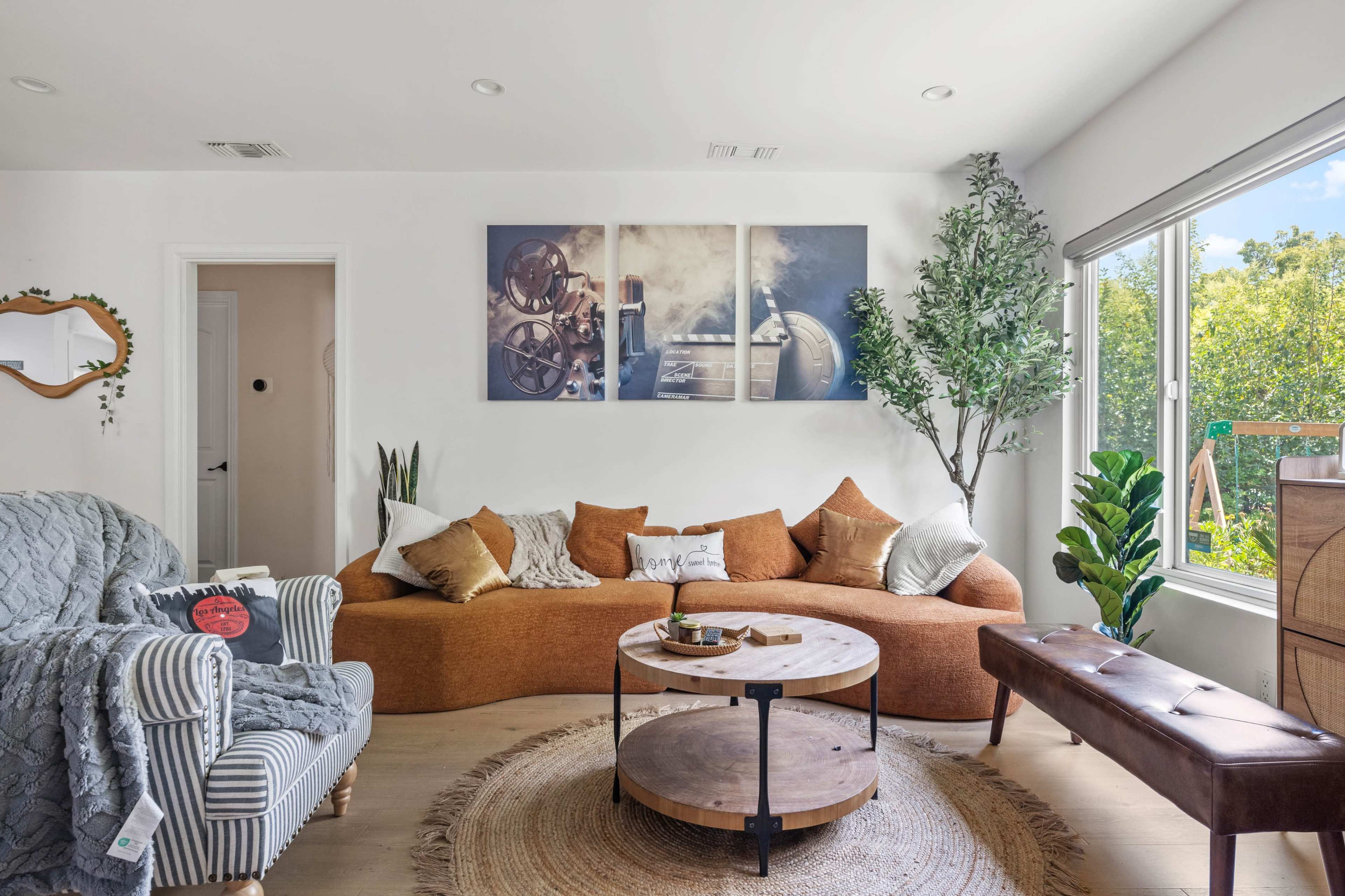 The image shows a modern living room with a curved brown sofa, a round wooden coffee table, and large windows that provide natural light and a view of greenery outside.