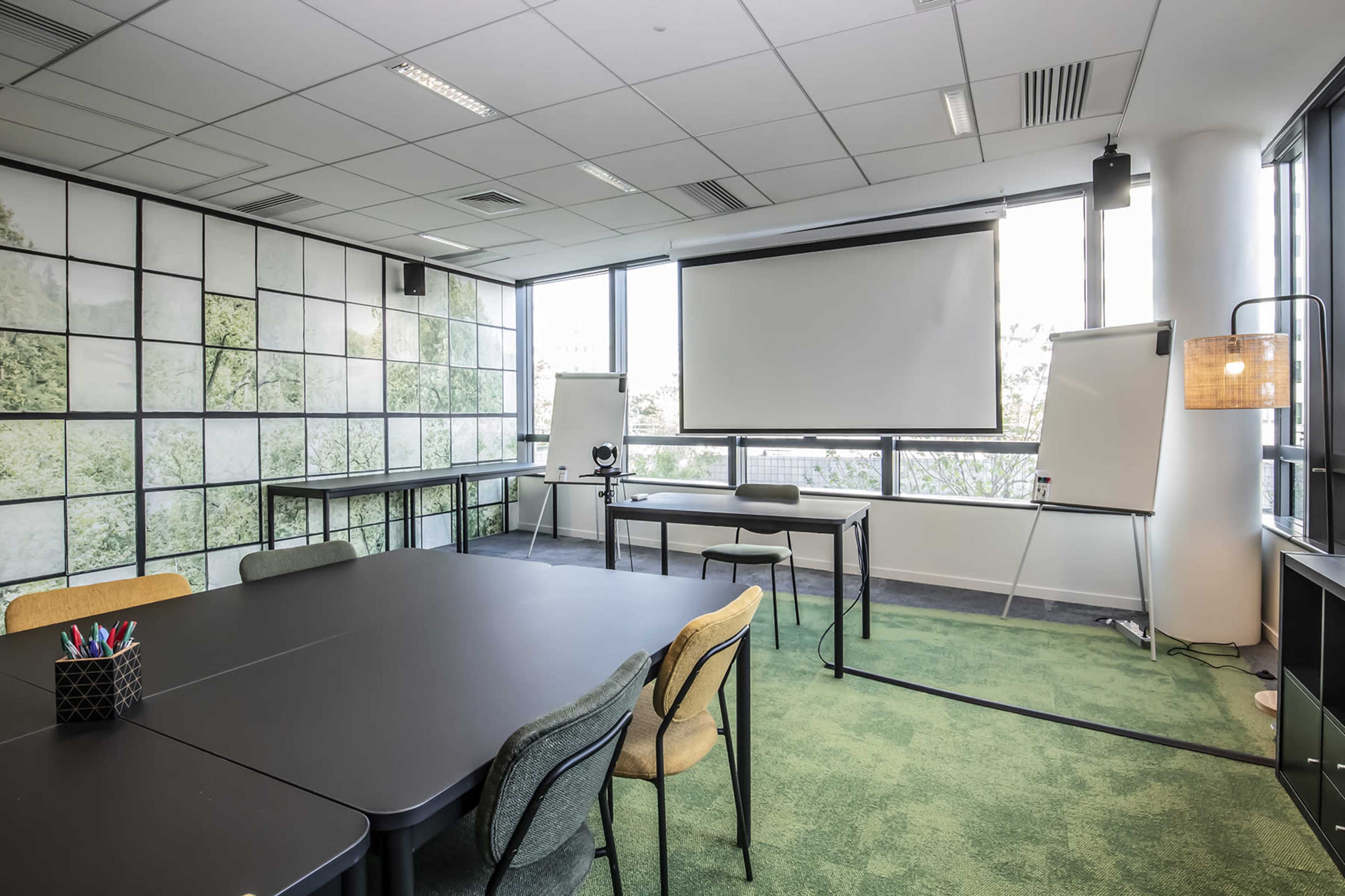A modern conference room features a large table surrounded by chairs, a projector screen, and whiteboards against a backdrop of green foliage on the walls.