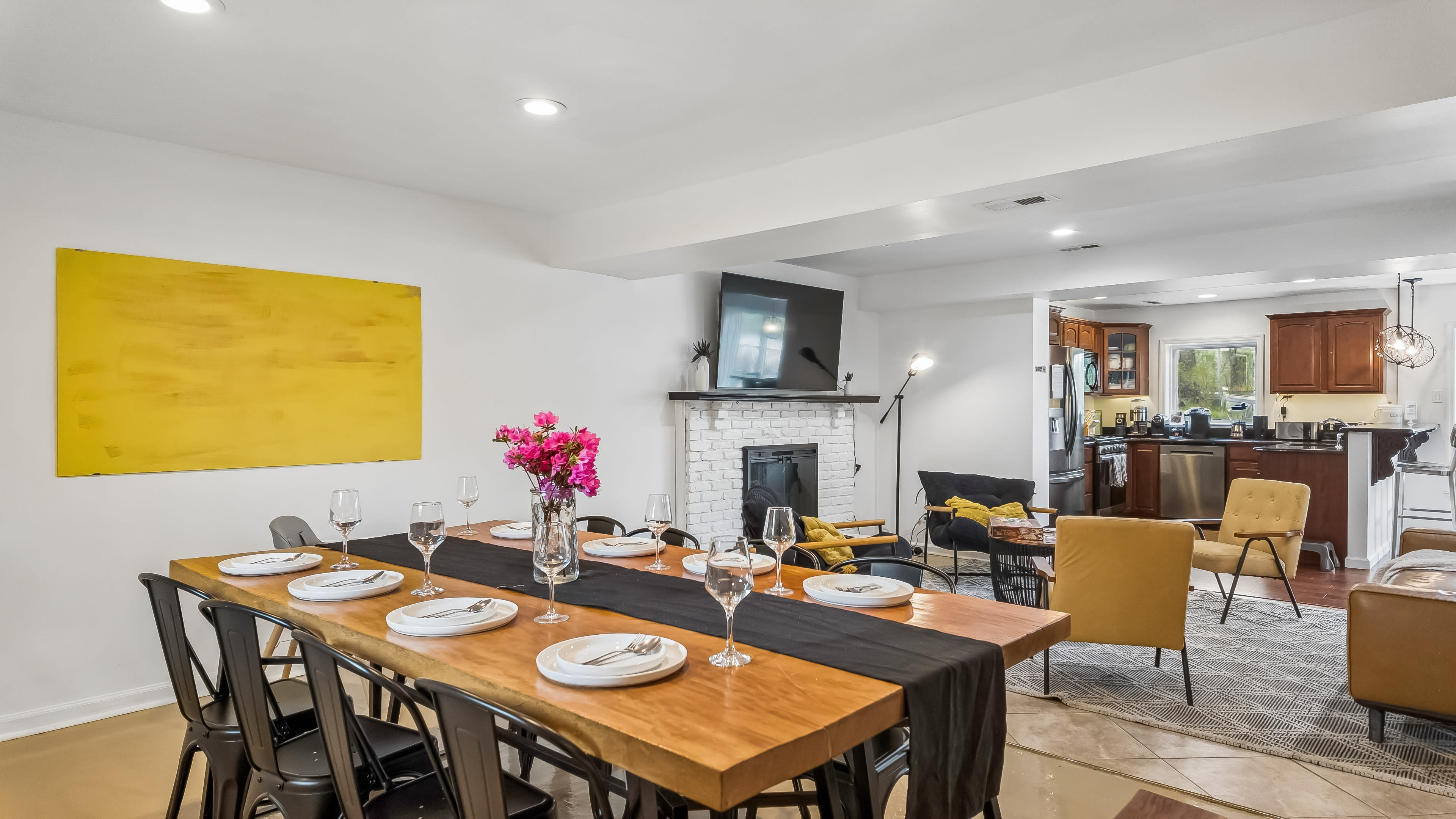 A dining area with a wooden table set for dinner, featuring a vase of flowers, and an adjacent living space with a fireplace and kitchen visible in the background.