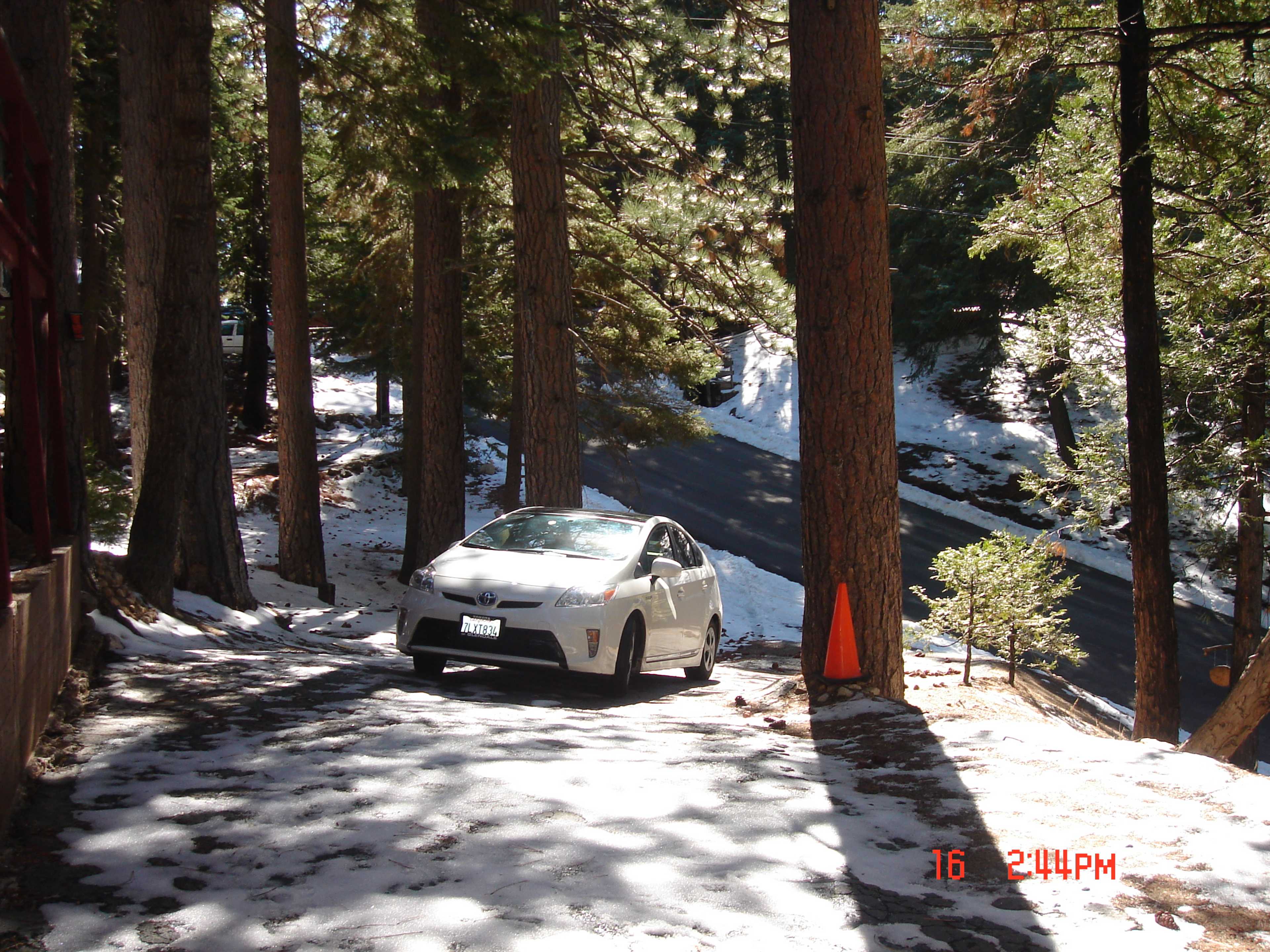 A white car is parked on a snowy road surrounded by tall trees.