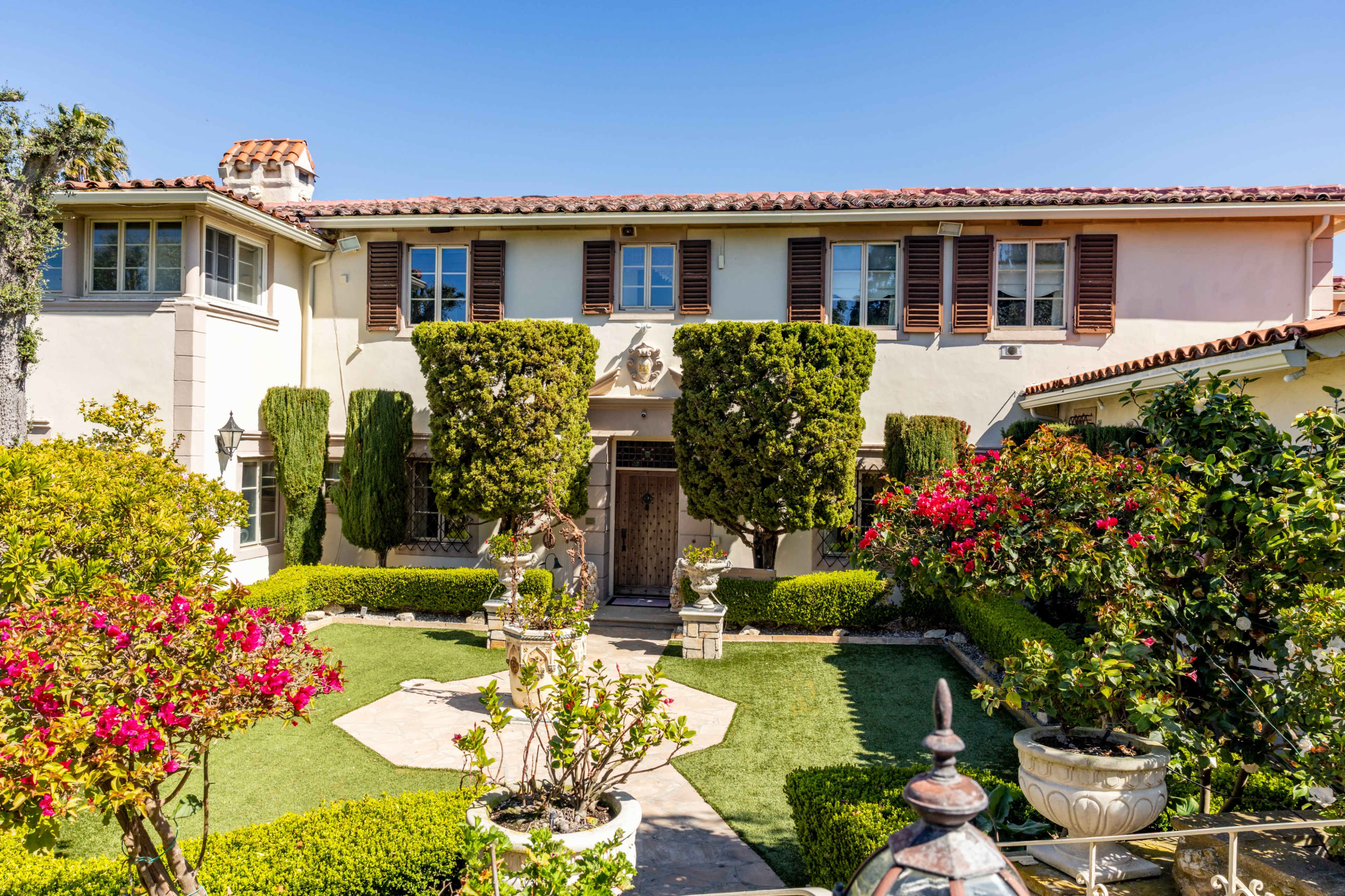 The image shows a two-story Mediterranean-style house with a tiled roof surrounded by well-maintained gardens and shrubs.