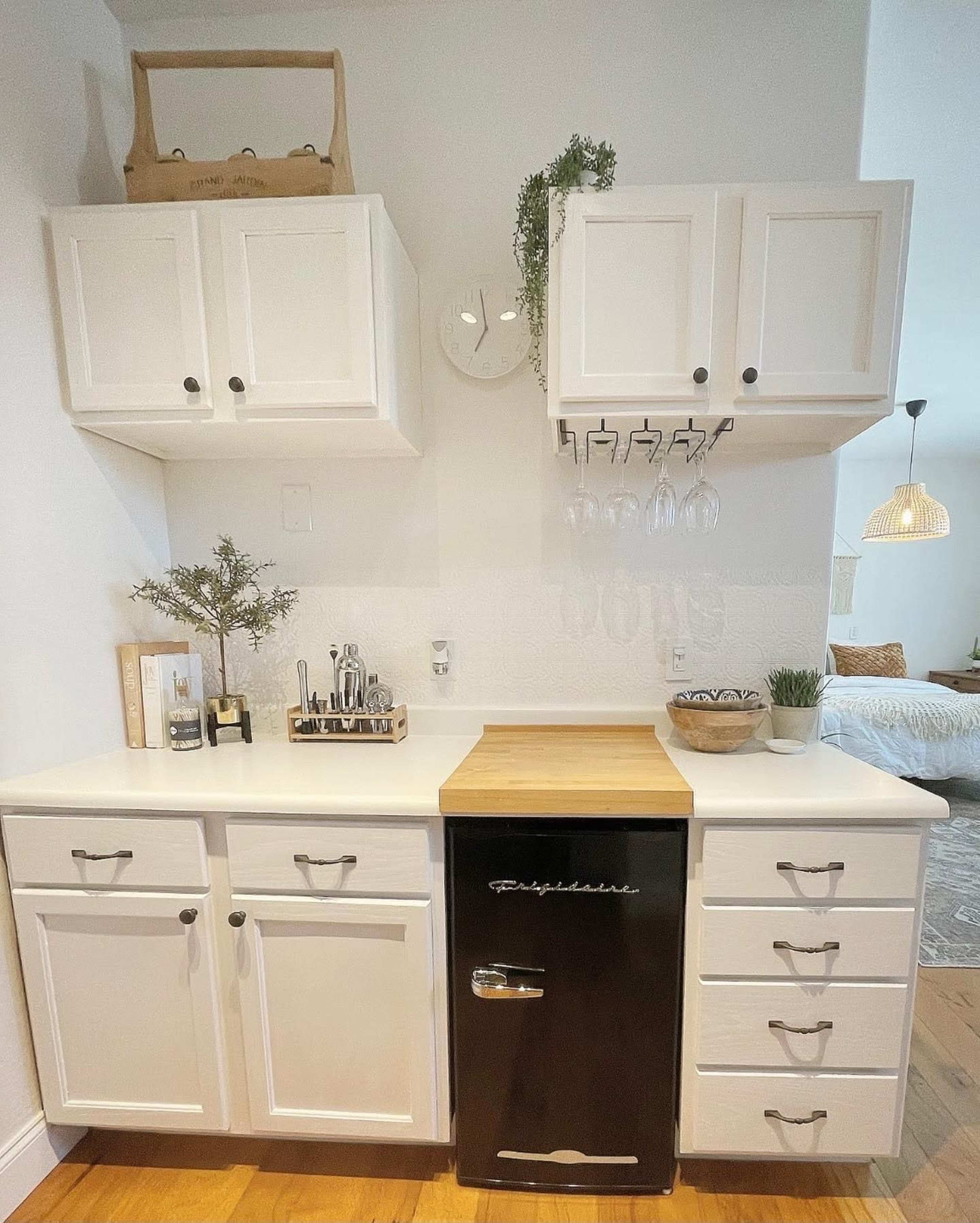 The image shows a compact kitchen area featuring white cabinets, a wooden countertop, and a small black refrigerator.