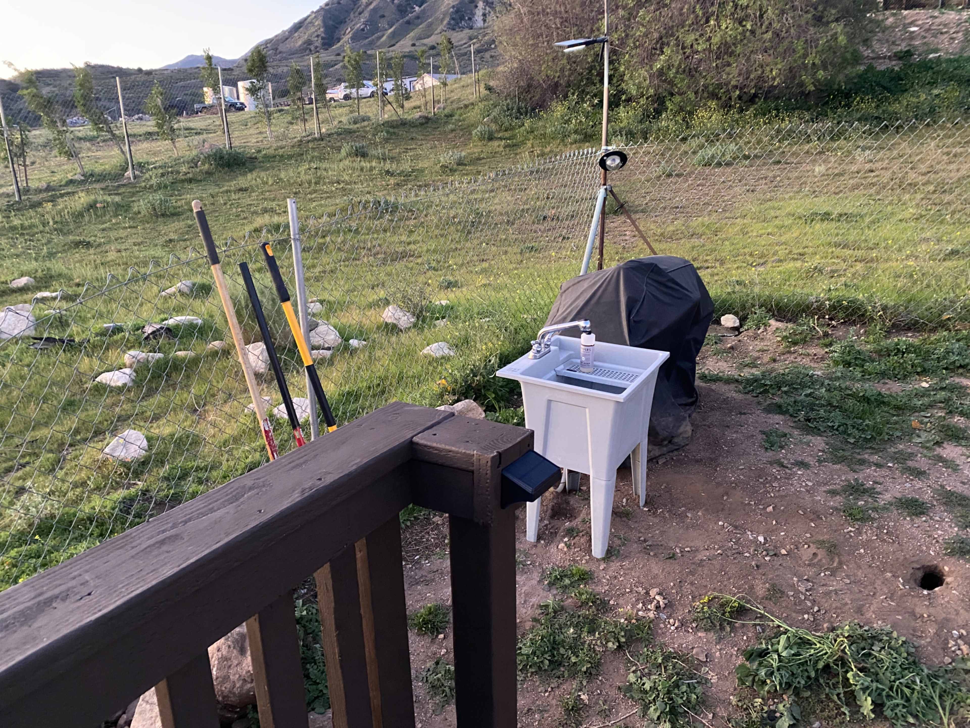 The image shows a fenced outdoor area with gardening tools leaning against a wooden railing and a covered grill on a small white table.