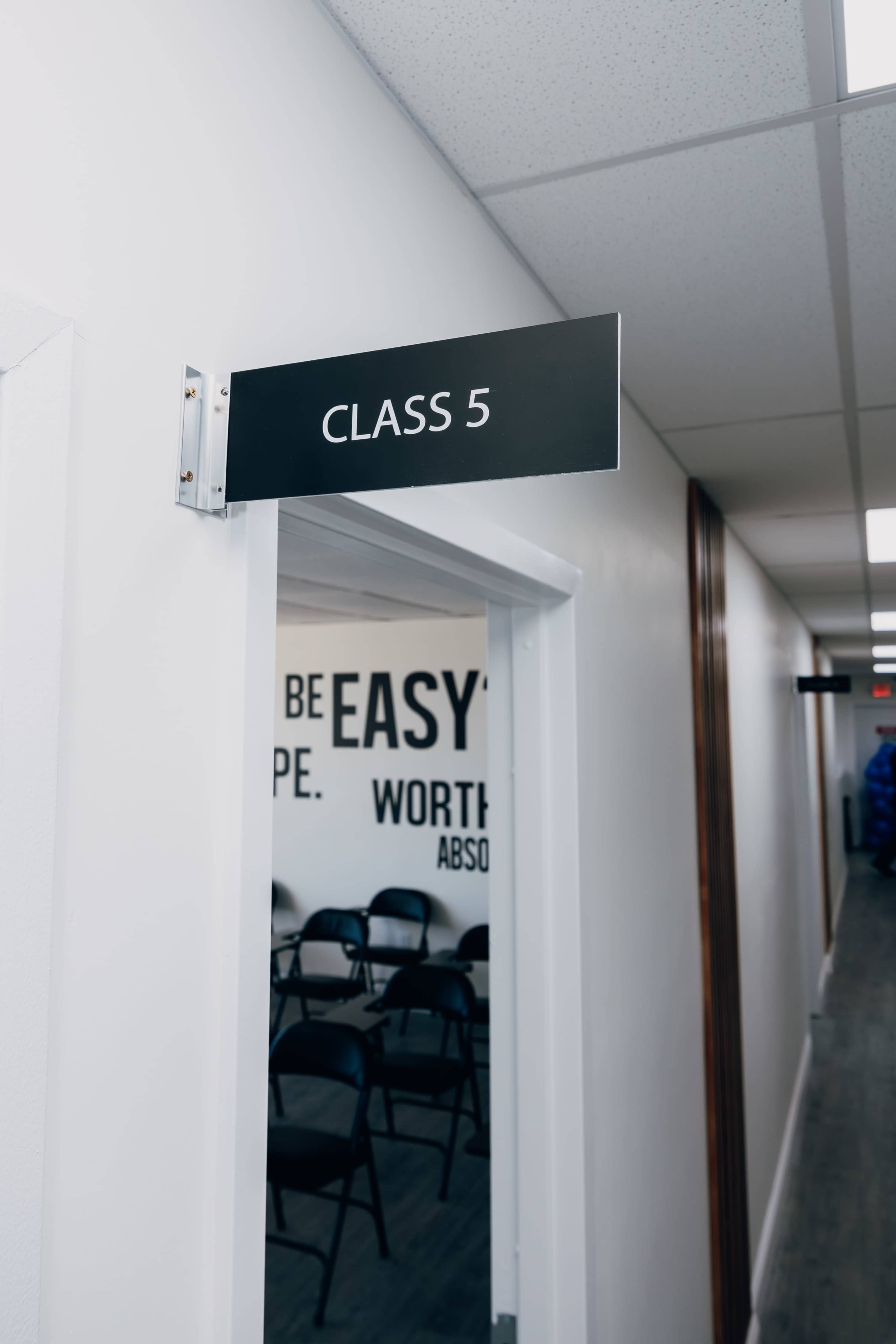 The image shows a hallway with a sign indicating "CLASS 5" on a door frame, leading to a classroom with several chairs inside.