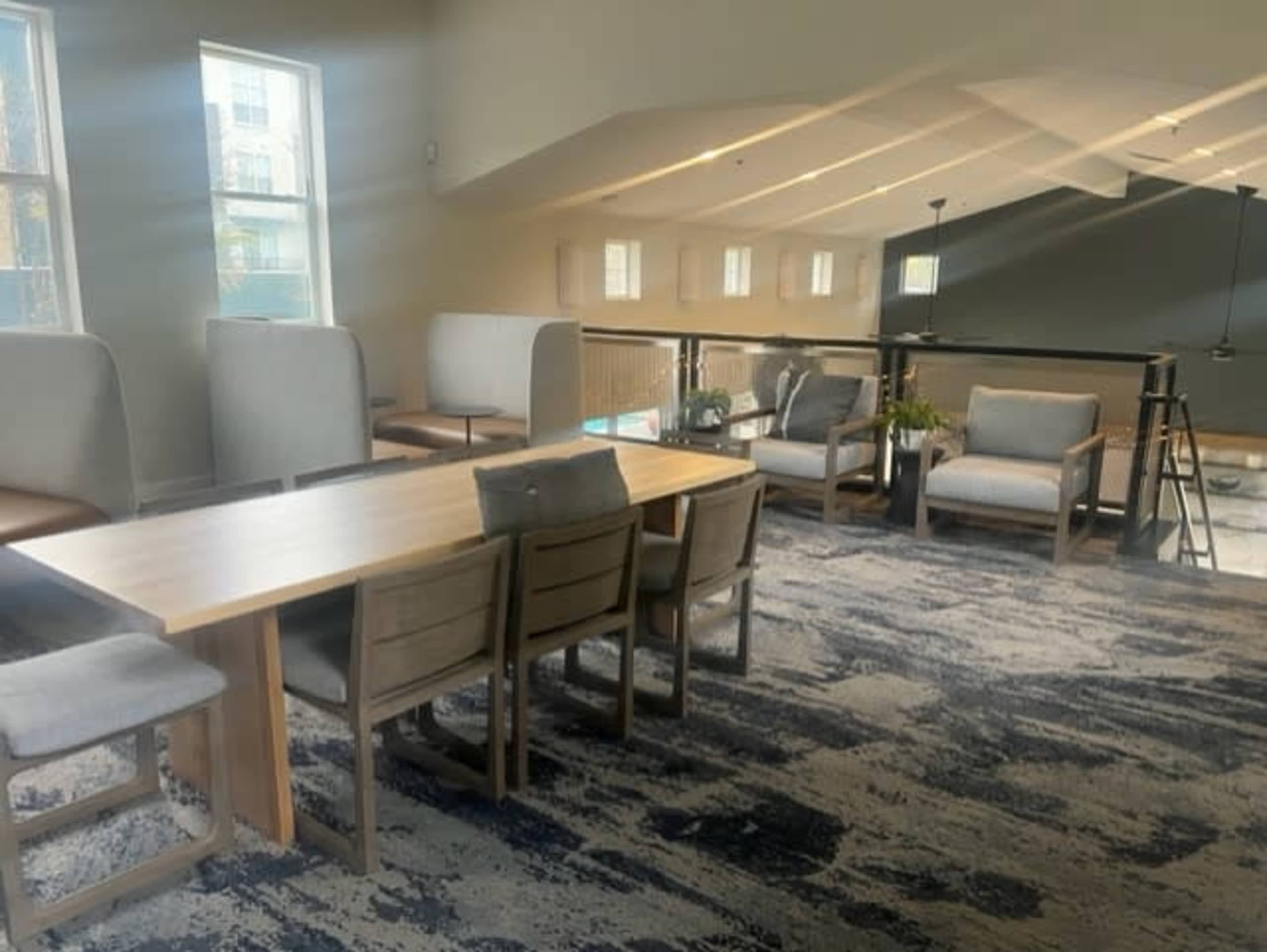 A modern lounge area with a long wooden table surrounded by chairs, set against a backdrop of light-colored furniture and large windows.